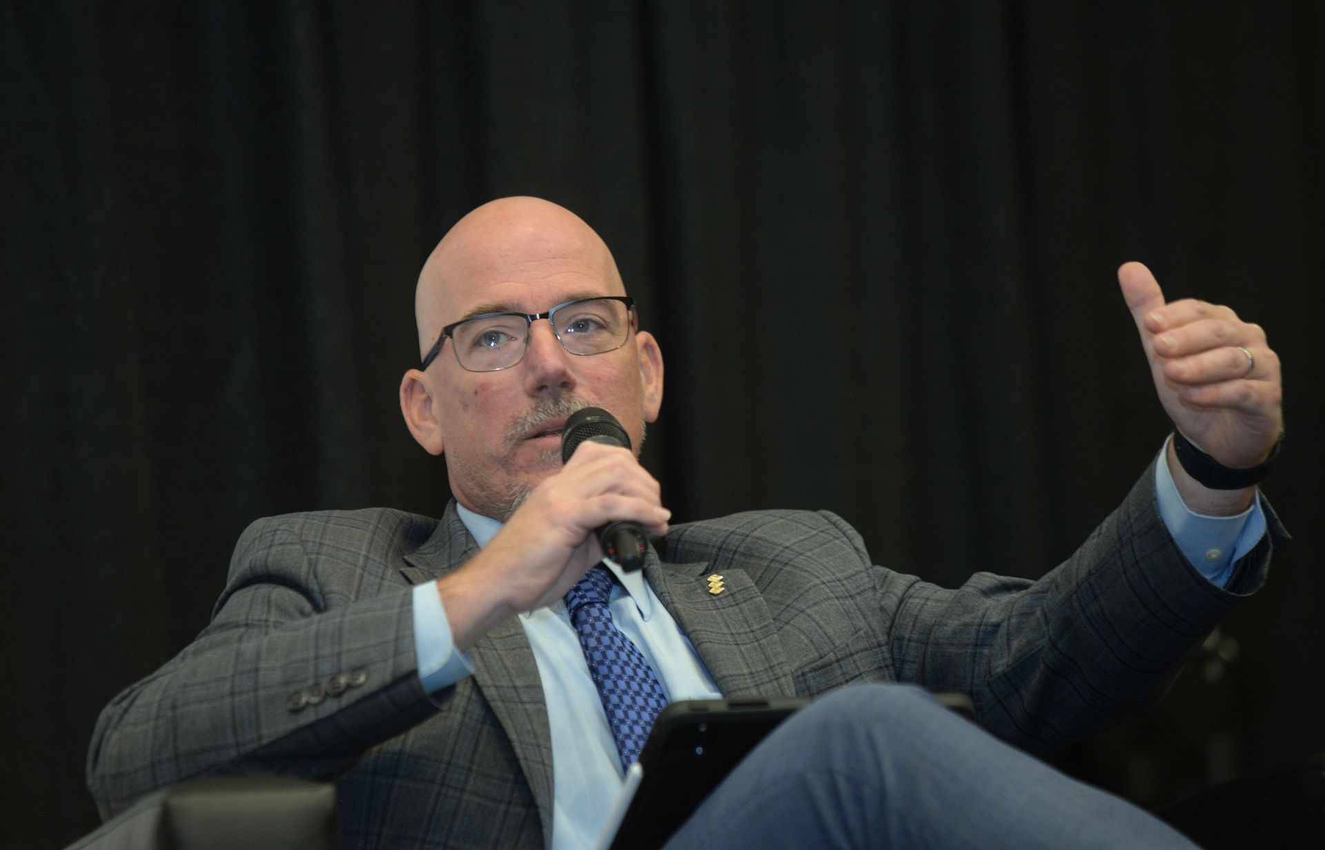Man with glasses speaks into a microphone, gesturing with his right hand.