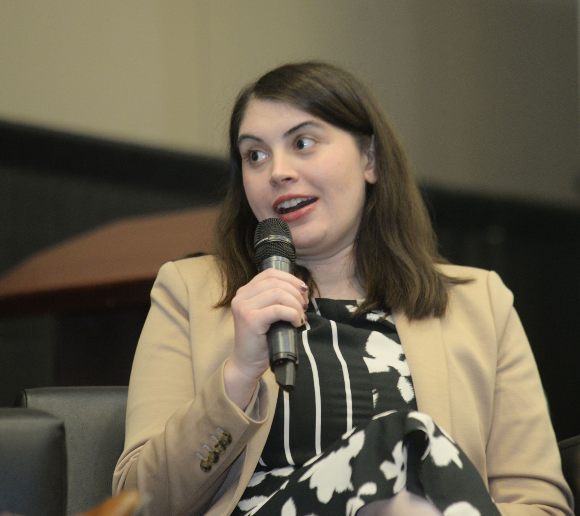 Woman speaking into a microphone, wearing a blazer and patterned dress, seated.