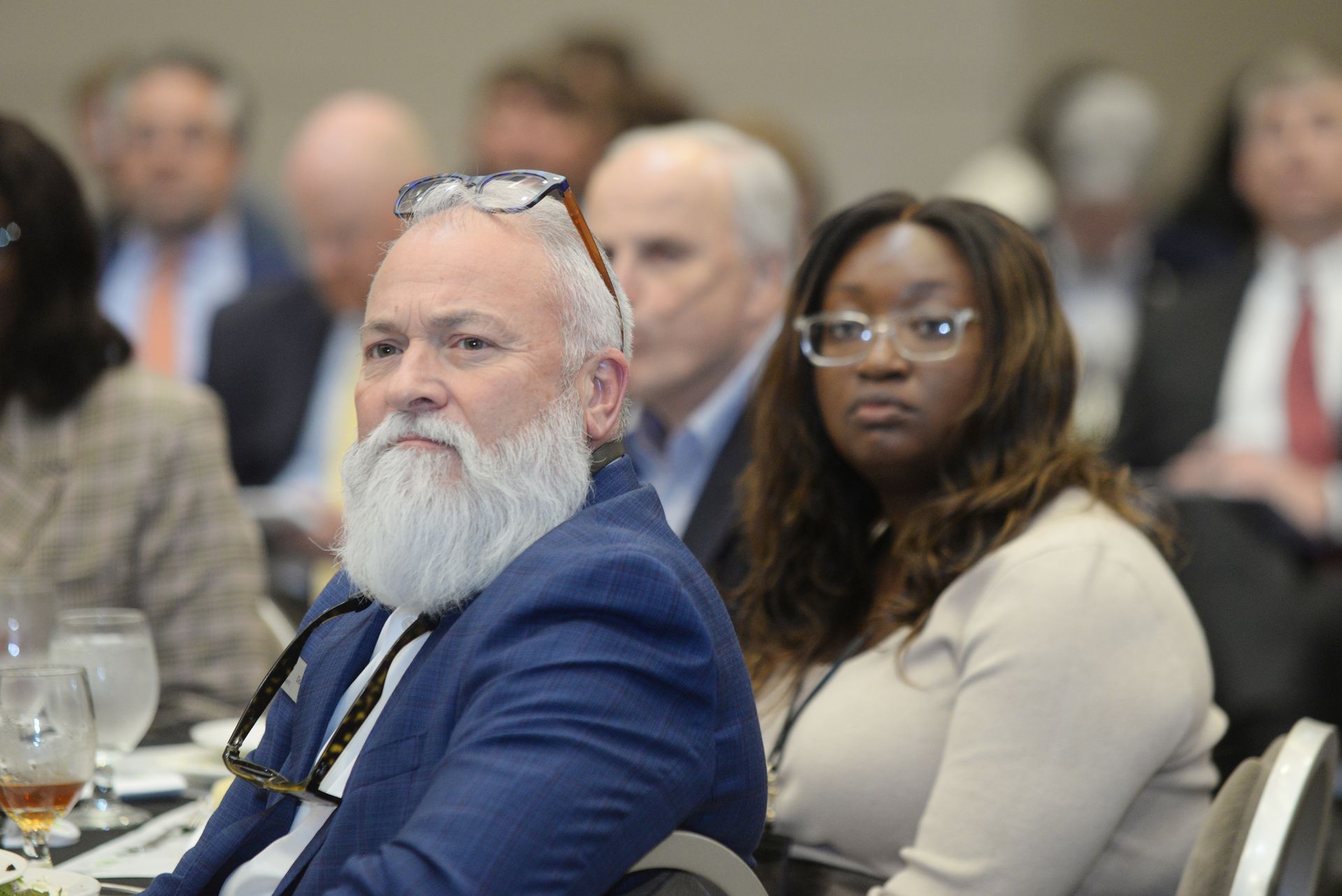 Attendees at a conference; man with a white beard and glasses, woman in glasses, and others in the background.