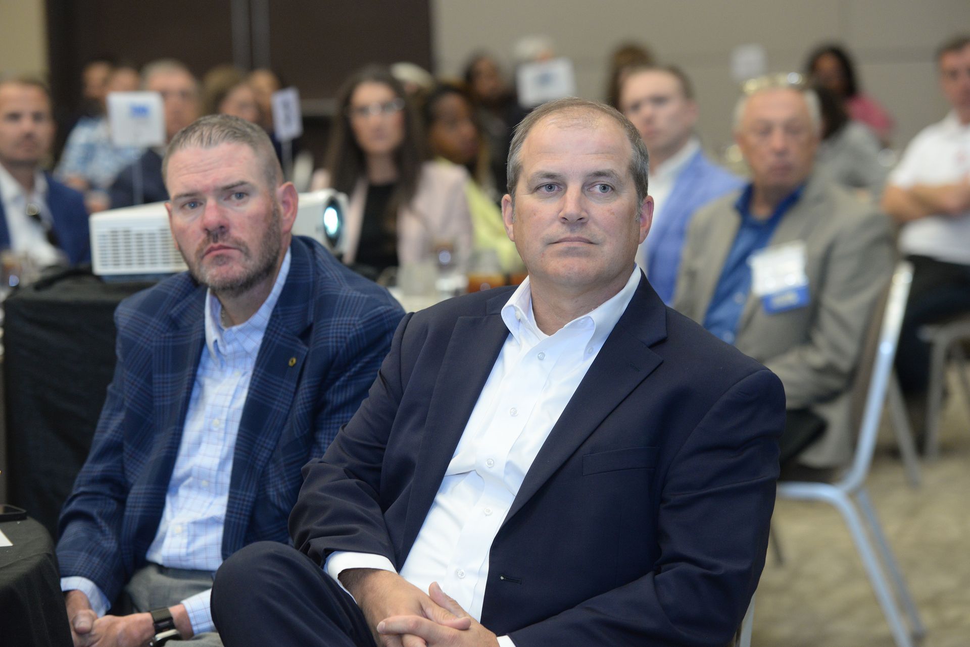 Two men in suits seated at a conference; other attendees in the background.