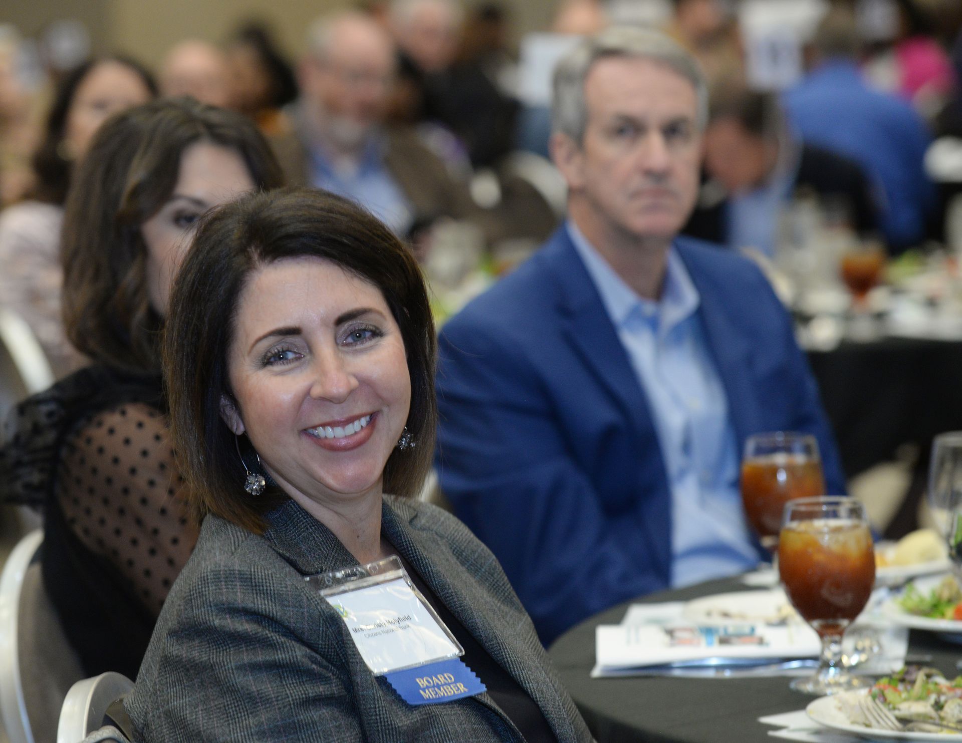 Woman in grey jacket smiles at camera during an event, man in blue blazer in background.
