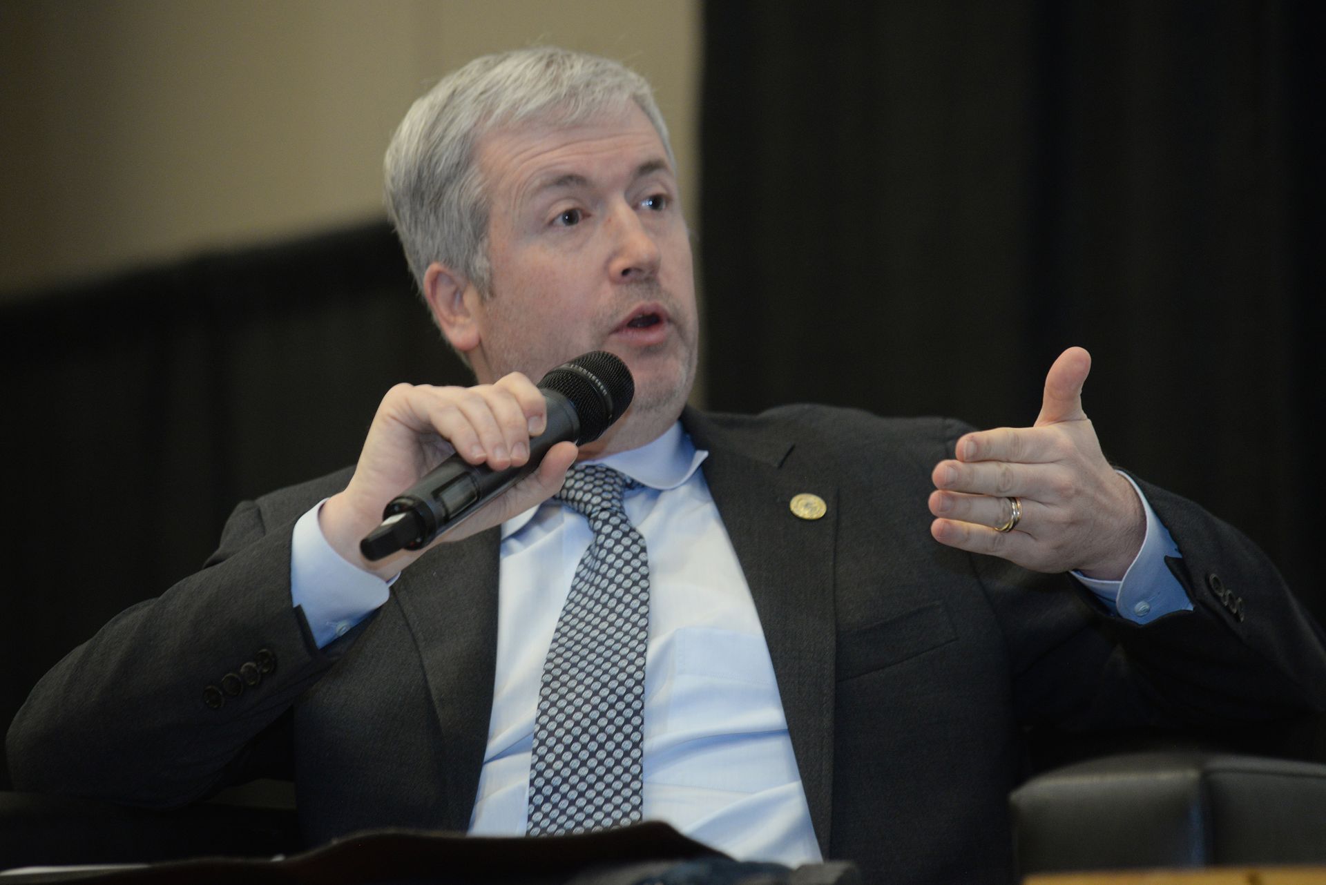 Man in suit speaks into microphone, gesturing with right hand at event.