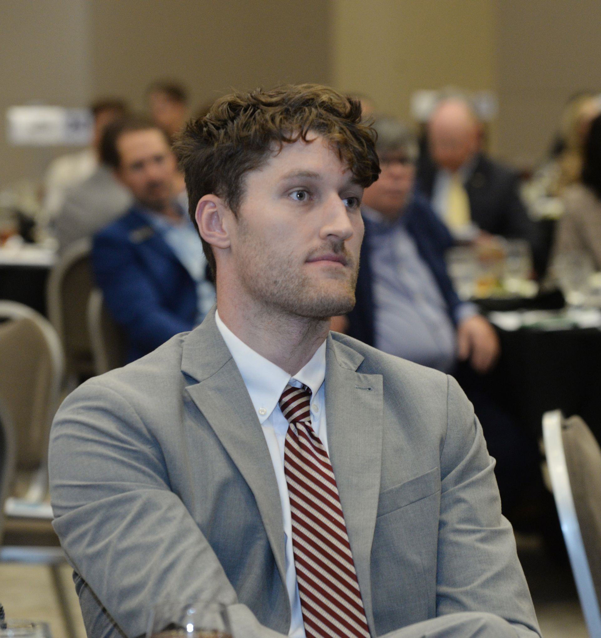 Man in a grey suit and striped tie sits at a conference table, looking to the side.