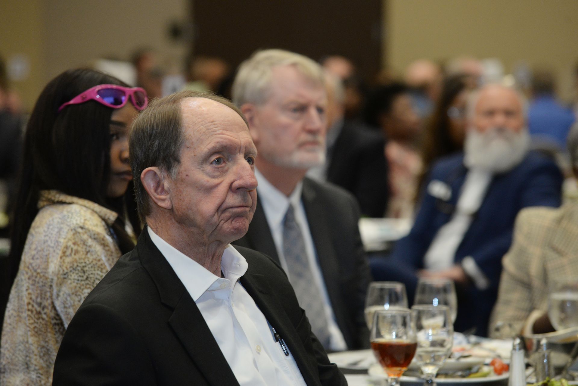 Attendees at a formal event; man in foreground with serious expression, others seated at tables.