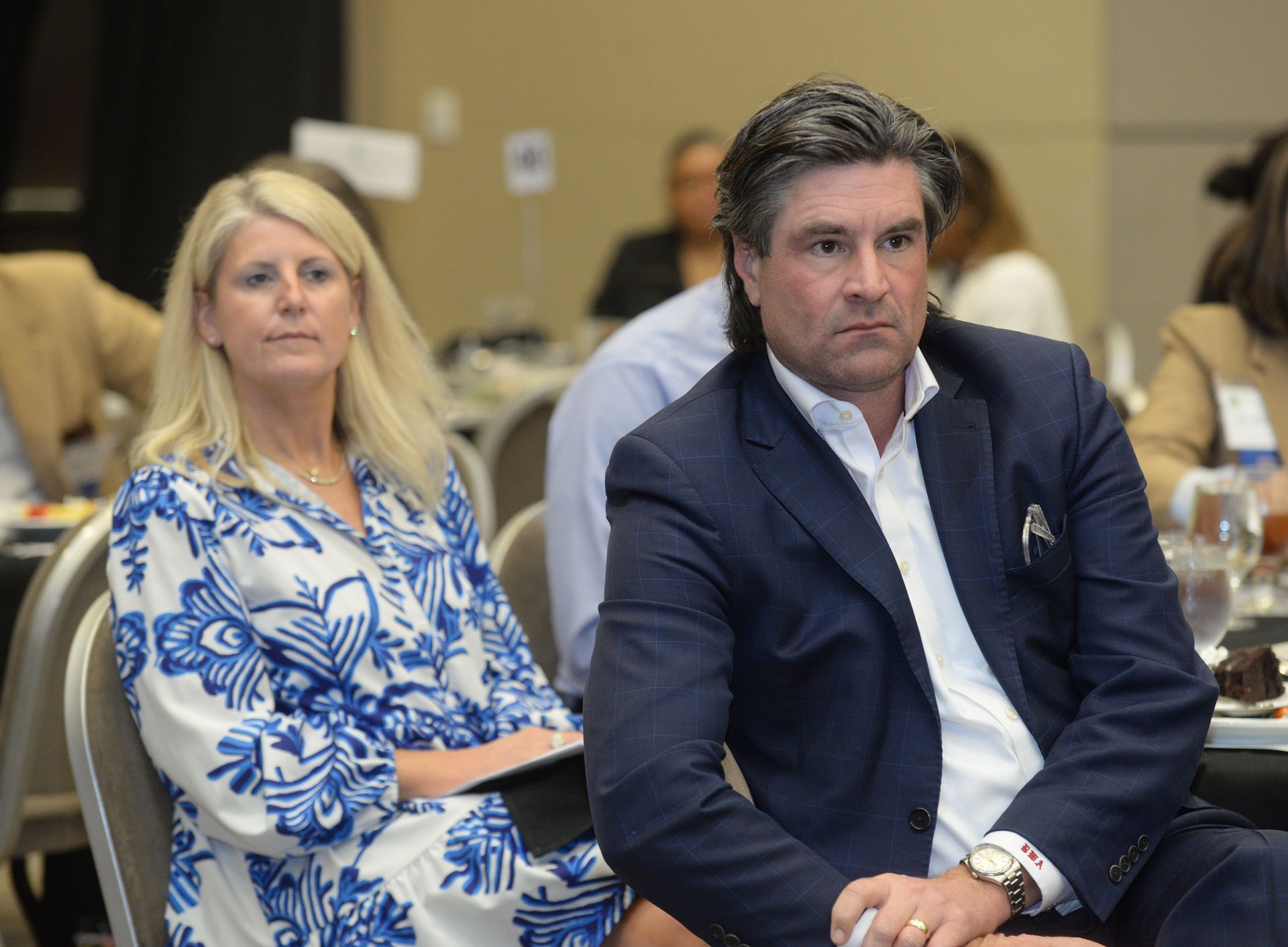 Woman in blue dress and man in suit at a conference, seated, listening.