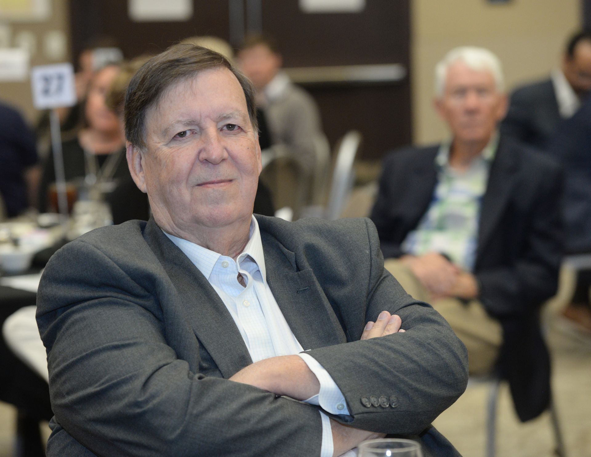 Man in a gray suit with arms crossed, seated at a table. Another man is visible in the background.