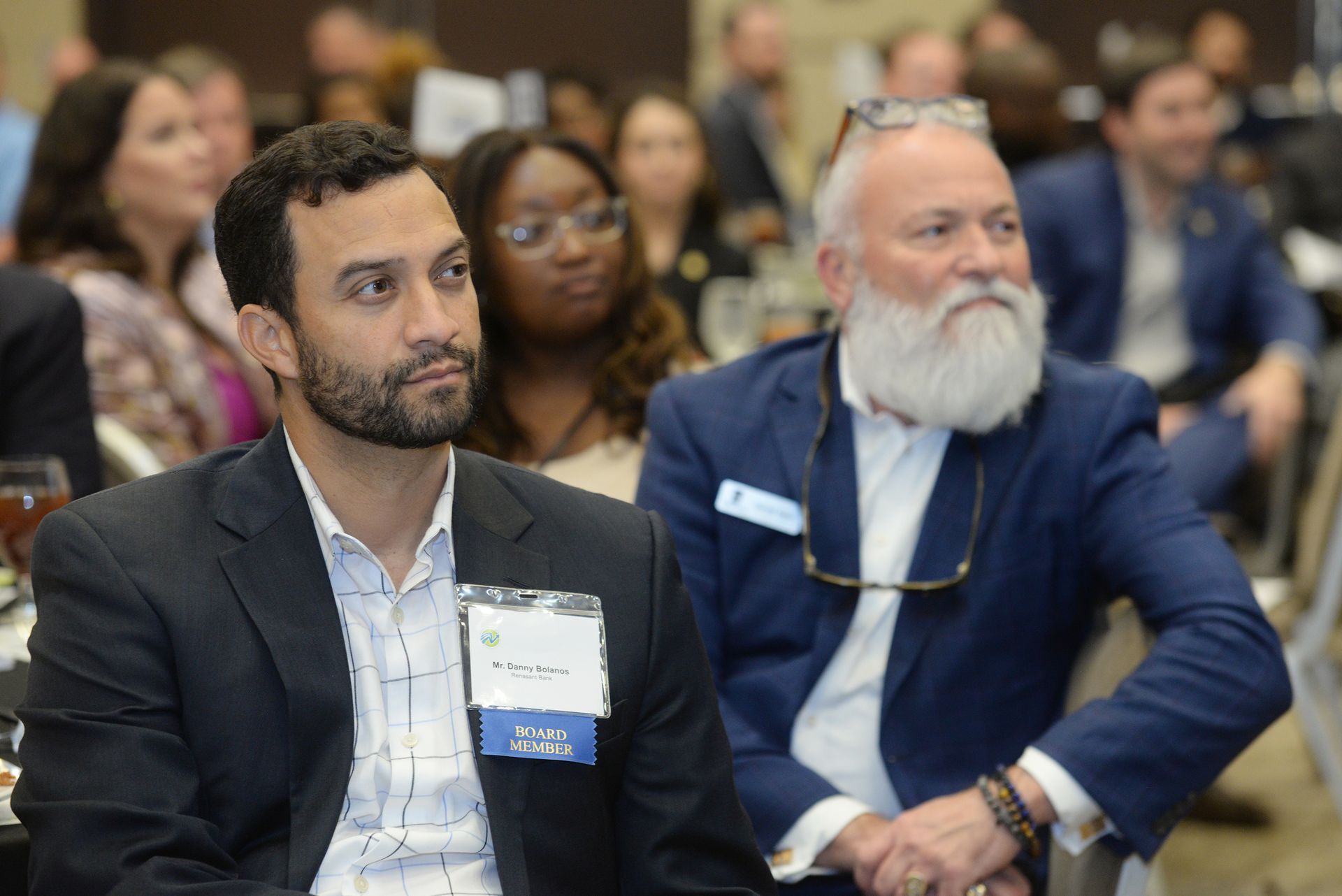 Two men in a conference setting; one with a beard and glasses, looking intently.