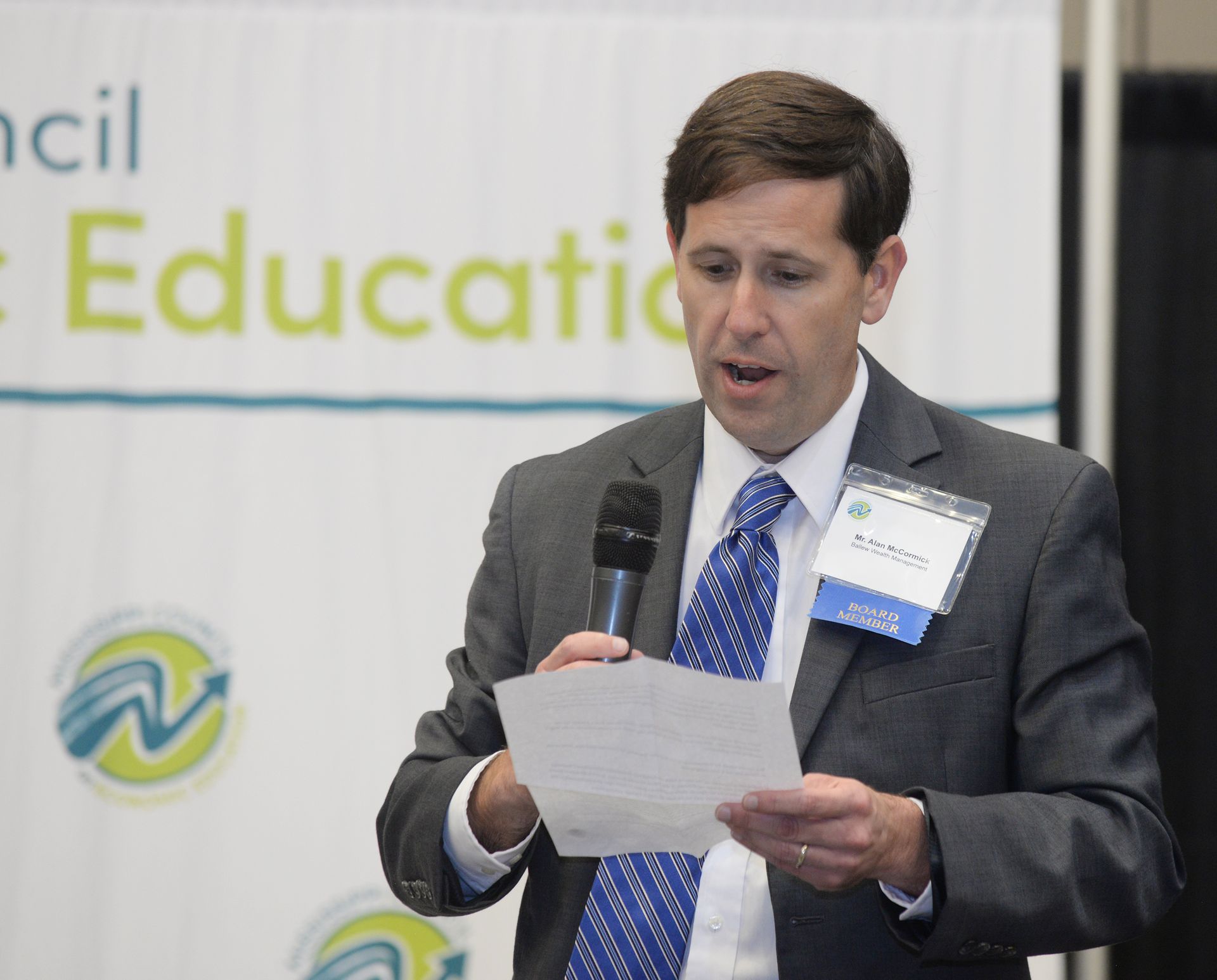 Man in suit speaking at a podium, holding papers and microphone, with event banner in background.
