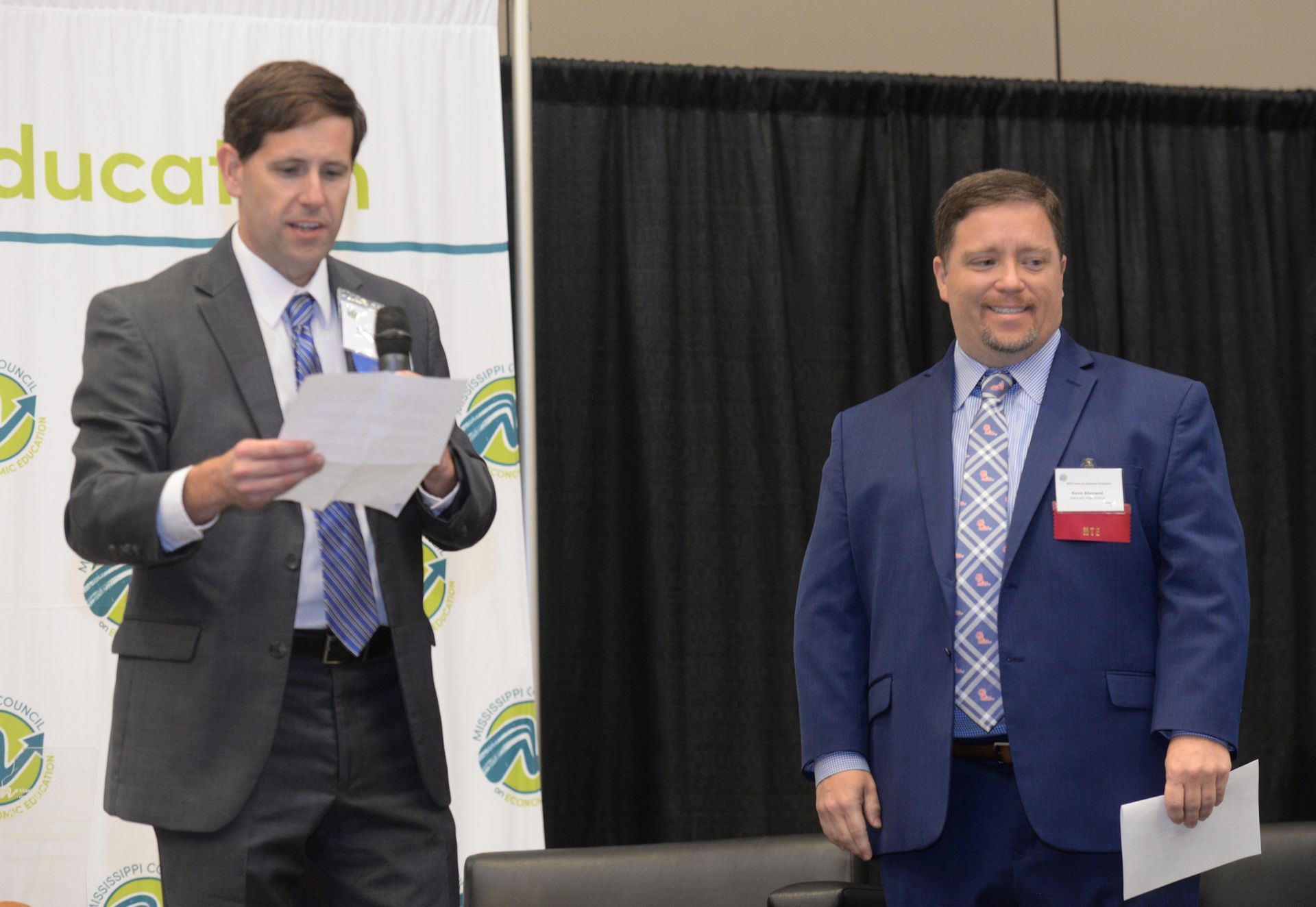 Two men in suits stand on a stage with a banner. One reads a paper, the other smiles.