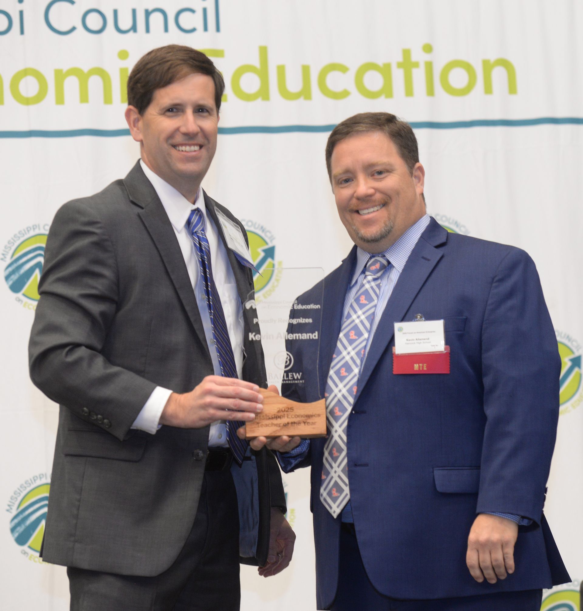 Two men in suits holding an award at an event with a backdrop.