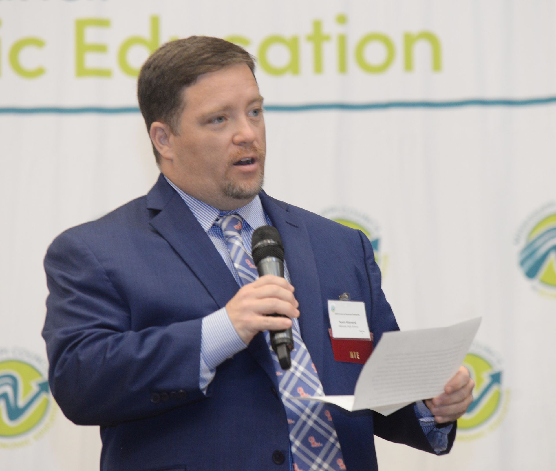 Man in a blue suit speaks into a microphone, holding papers. He appears to be giving a presentation at an event.