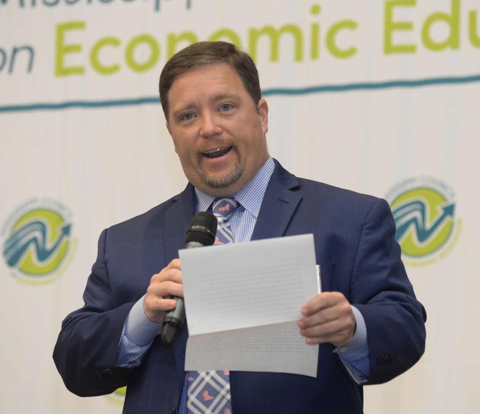 Man in blue suit speaking into microphone, holding paper. Background reads 