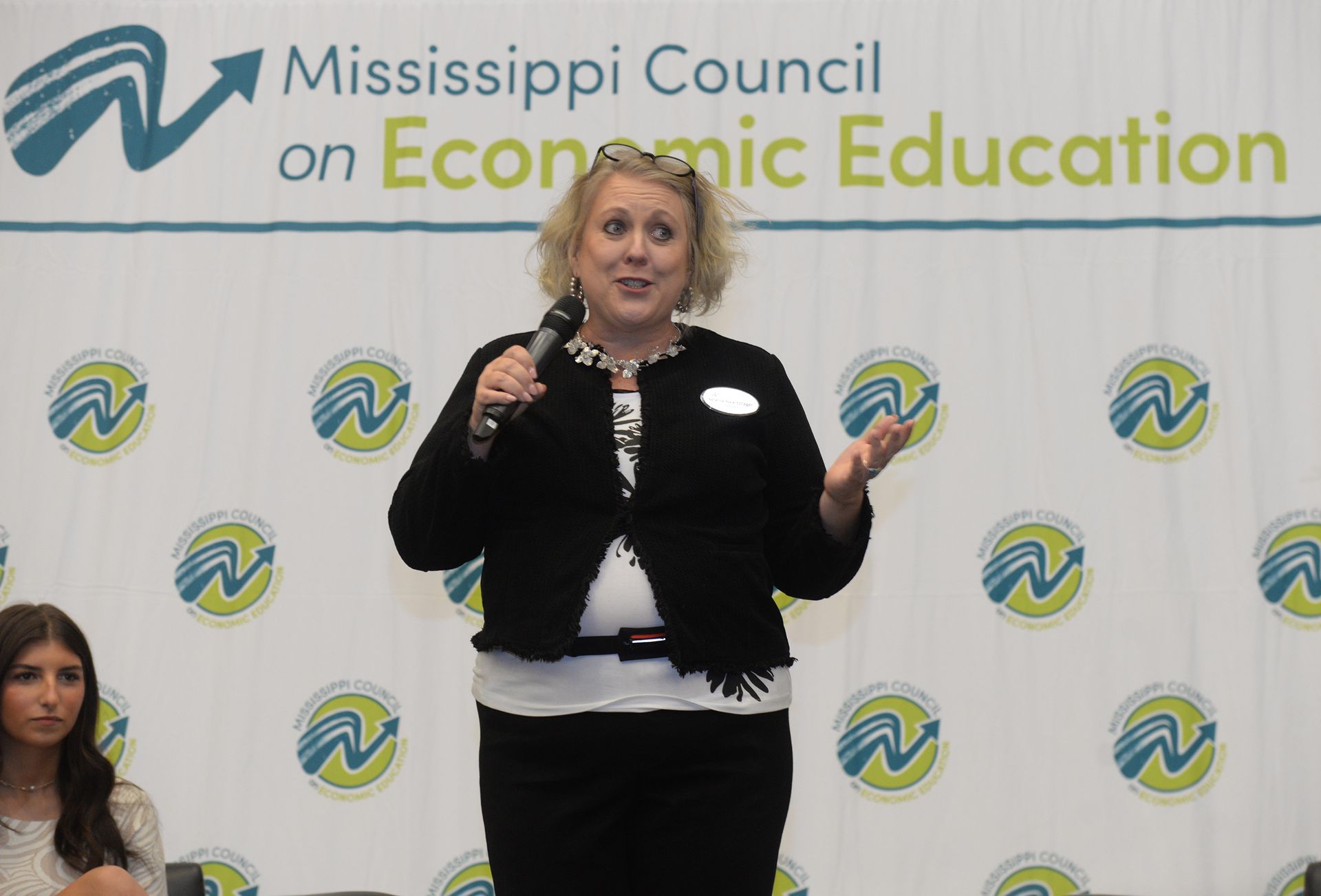 Woman speaking at podium, arms raised, in front of a backdrop with 