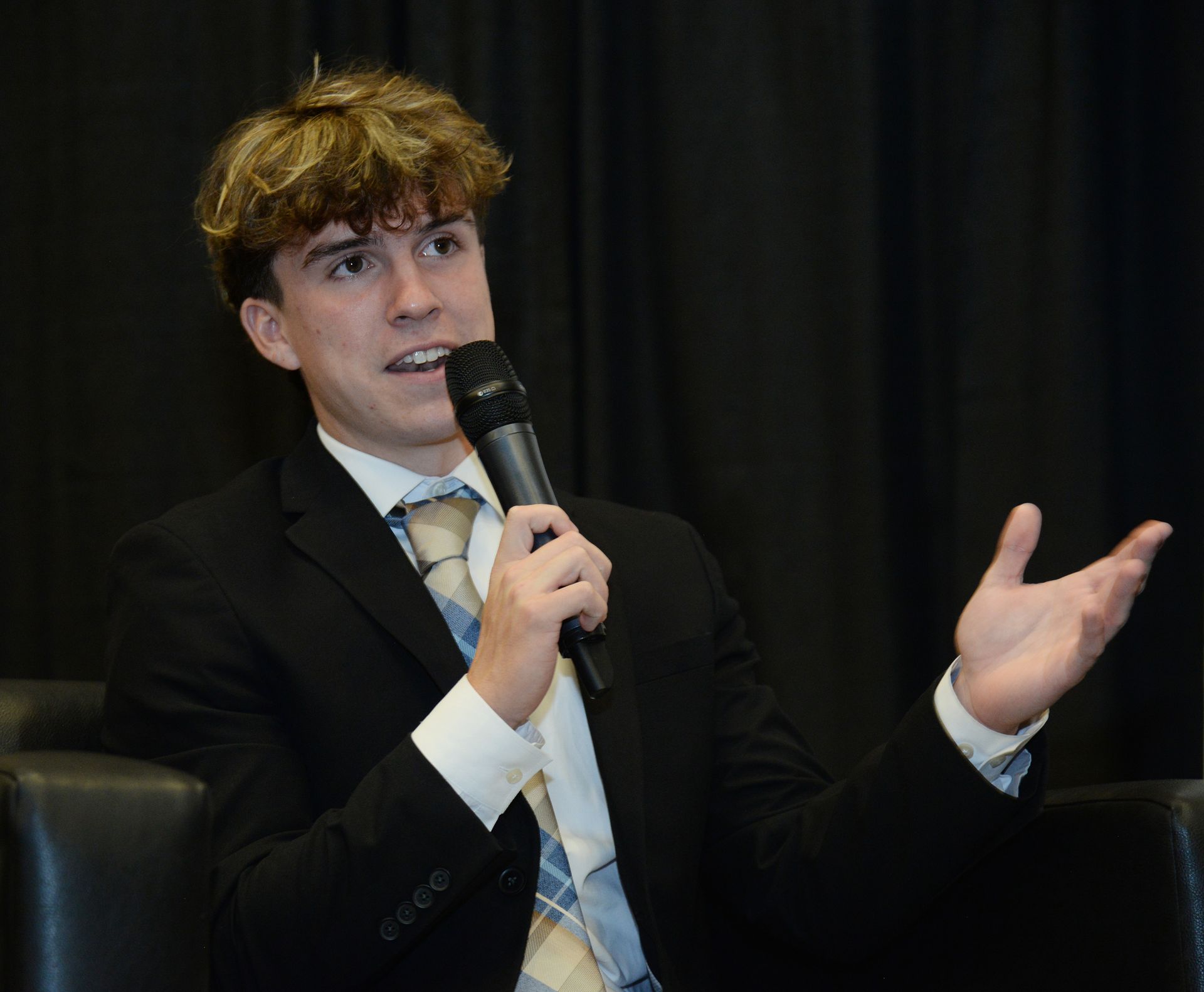 A young man in a suit speaks into a microphone, gesturing with his right hand.