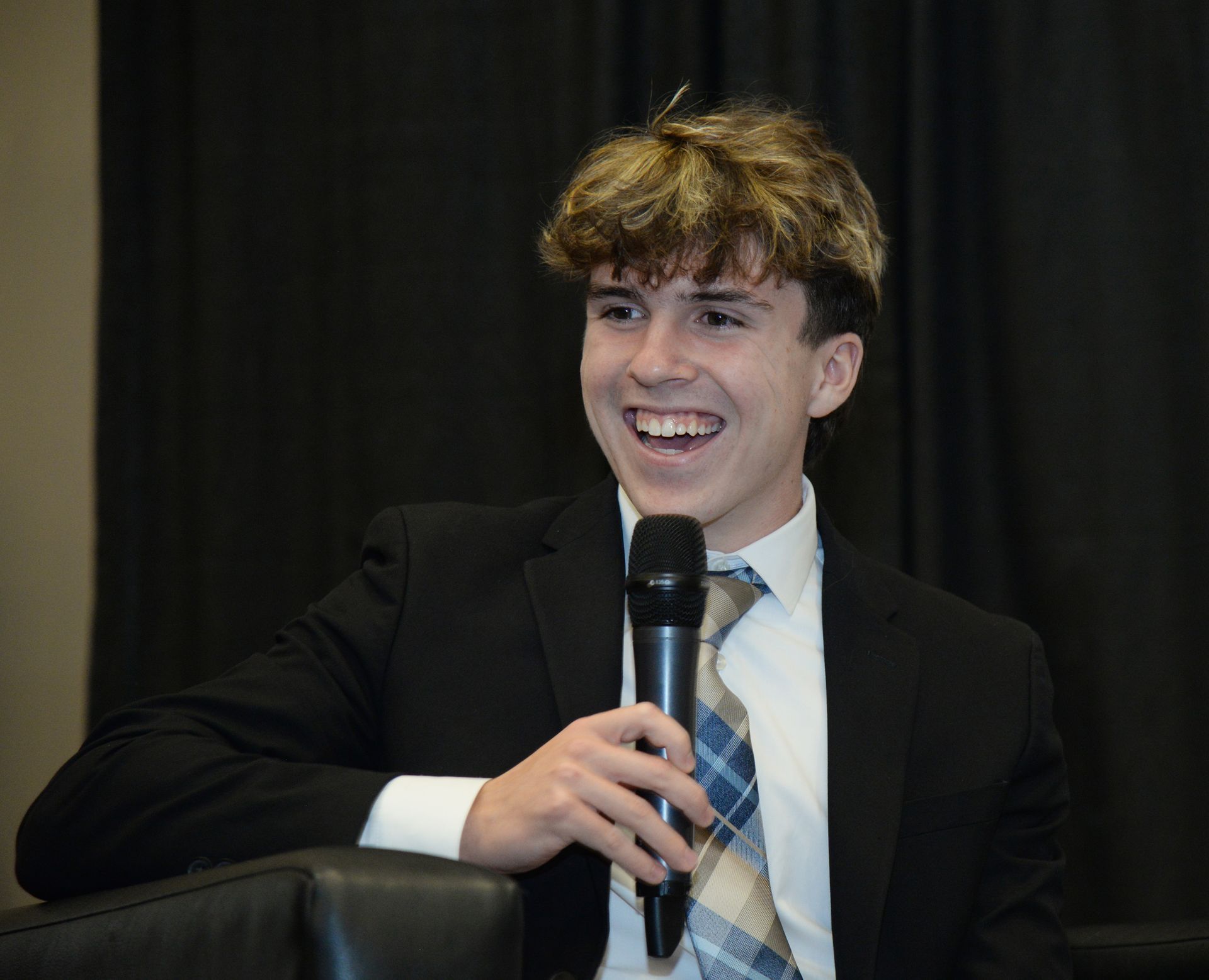 Young man in a suit smiles, holding a microphone, seated. Black backdrop.