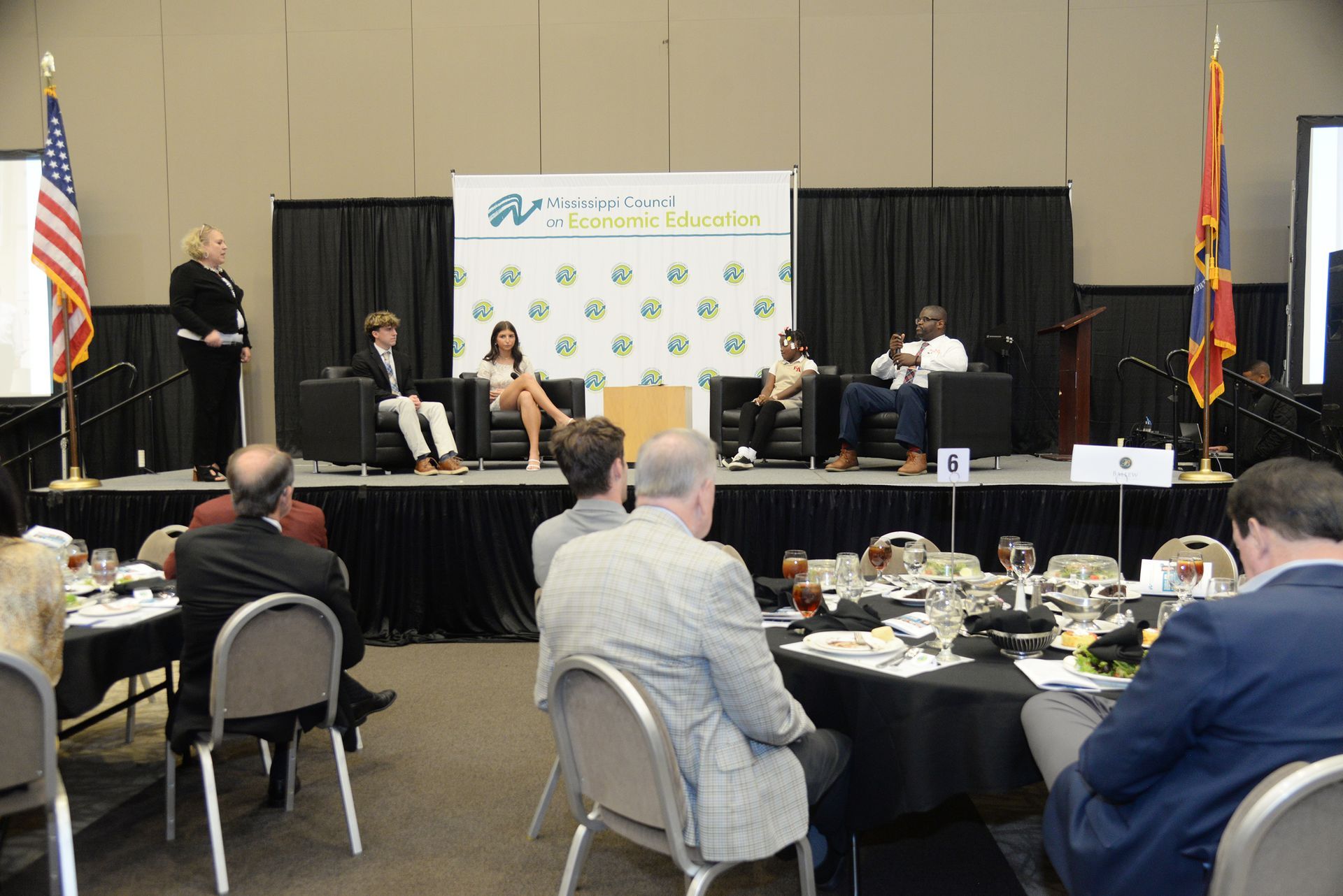 Panel discussion on stage with seated speakers, audience at tables, flags, and a banner.