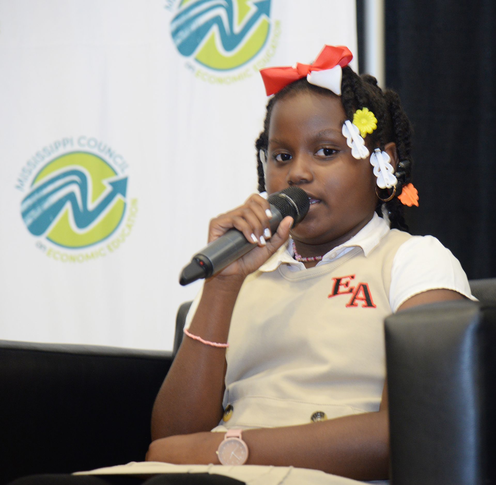 Girl in school uniform speaking into a microphone at a conference; logo in background.