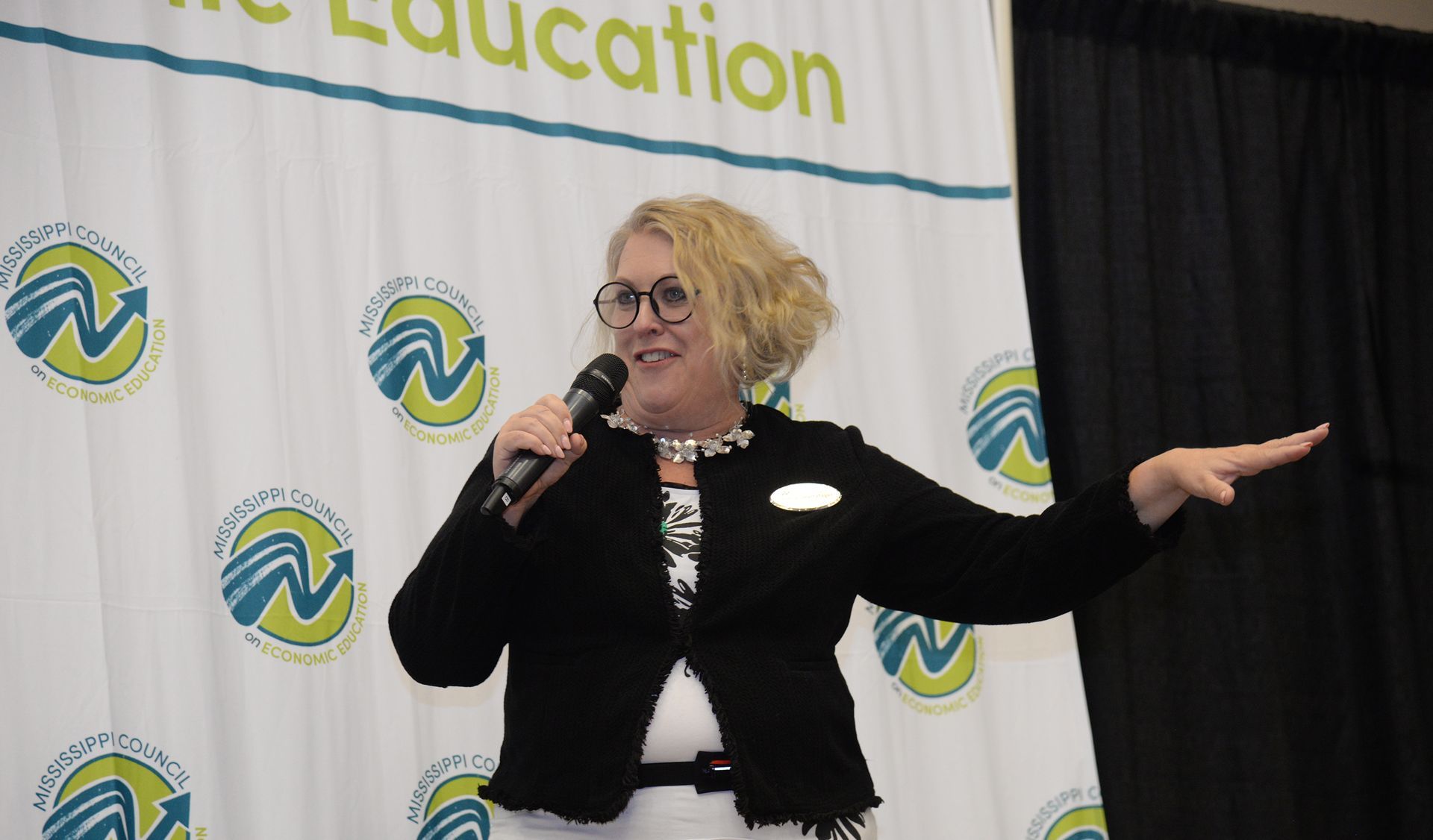 Woman speaking into a microphone, gesturing with her right hand. She's in front of a banner with a logo.