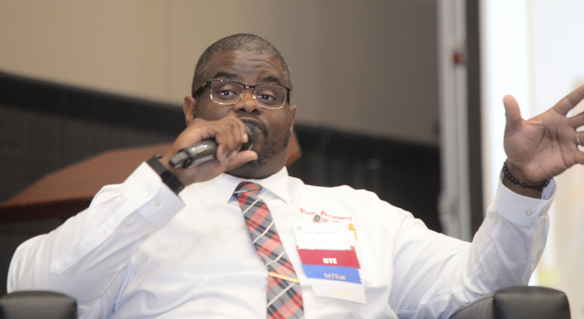 Man in glasses, holding microphone, gesturing with right hand, speaking at conference.