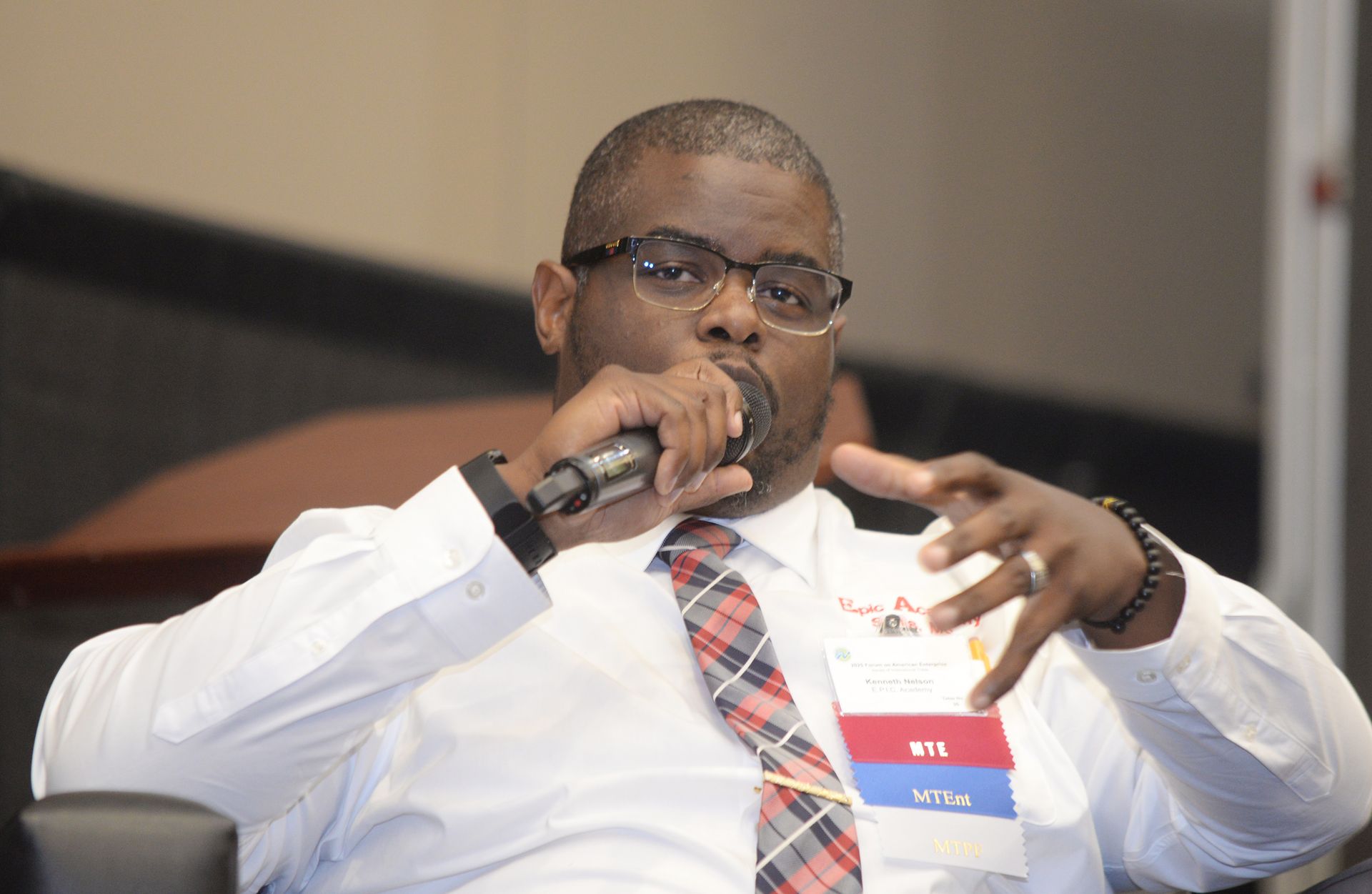 Man speaking into a microphone, gesturing with his hands. He's wearing glasses, a shirt, and a tie, seated indoors.