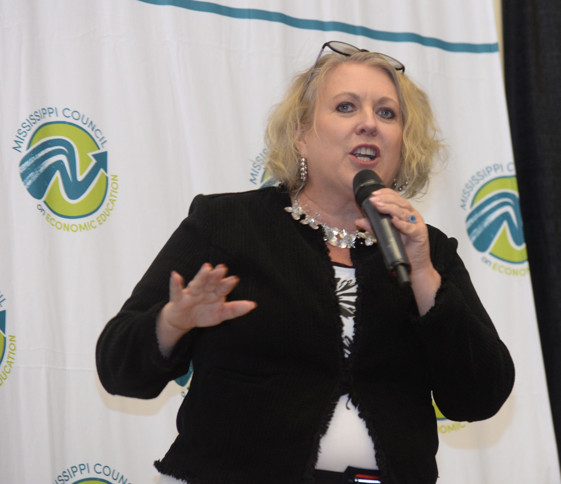 Woman speaking into microphone at an event, gesturing with her hands. Backdrop with a logo.