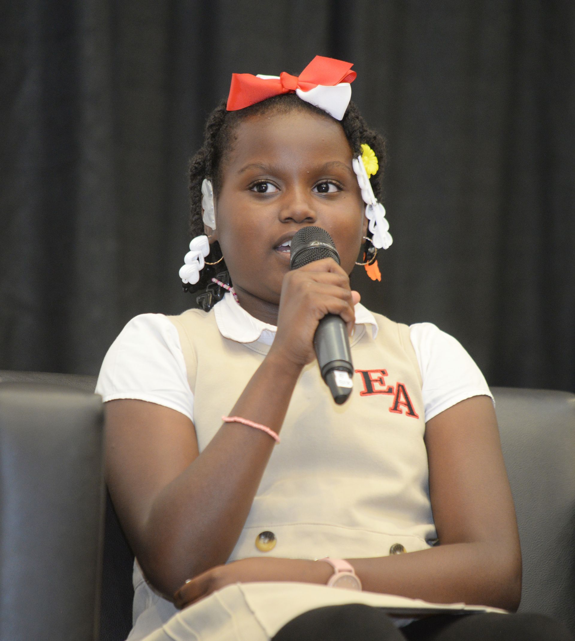Girl in school uniform speaking into a microphone, sitting down, wearing a bow.