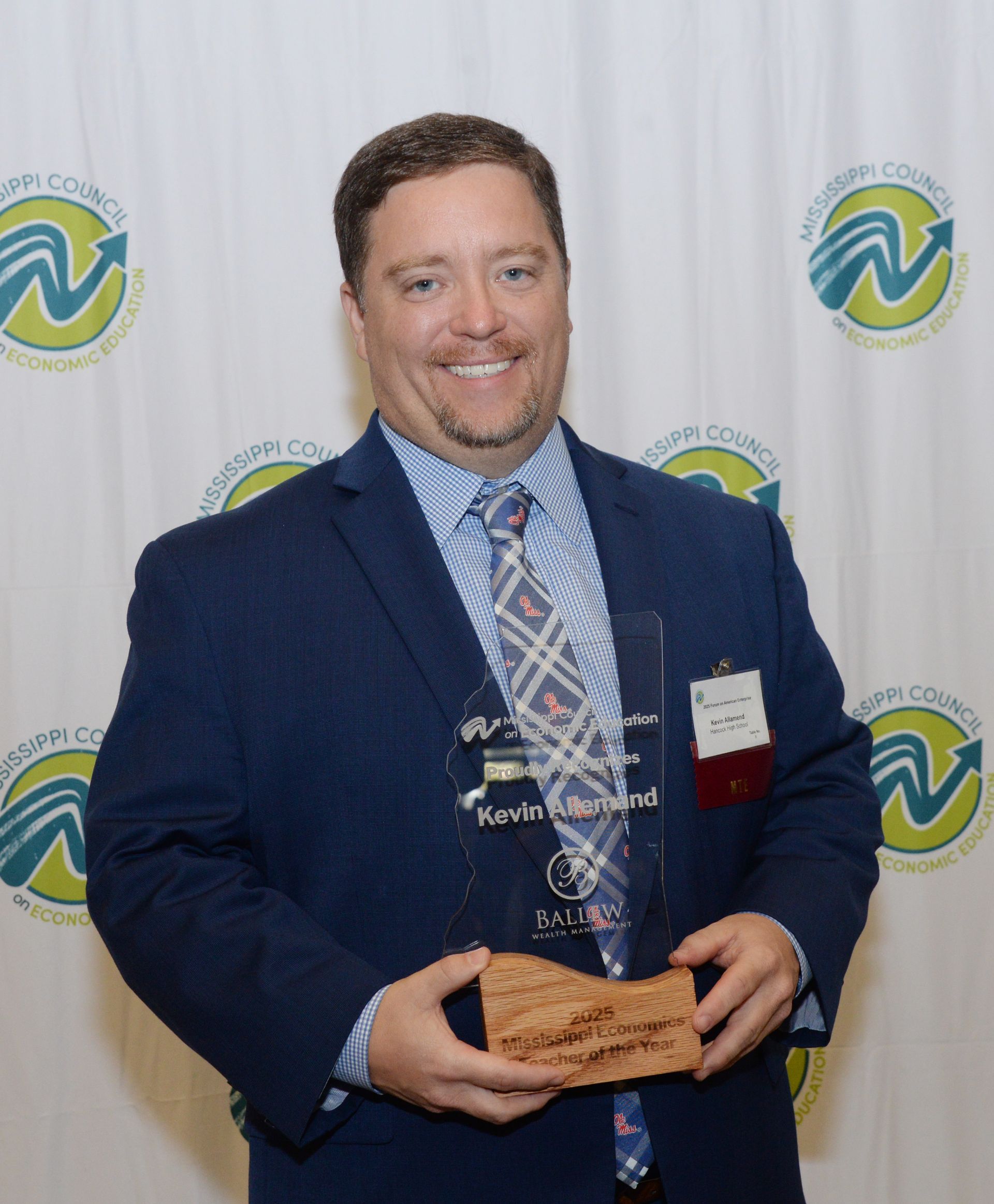 Man in a blue suit holds an award; smiling at the camera, event backdrop.