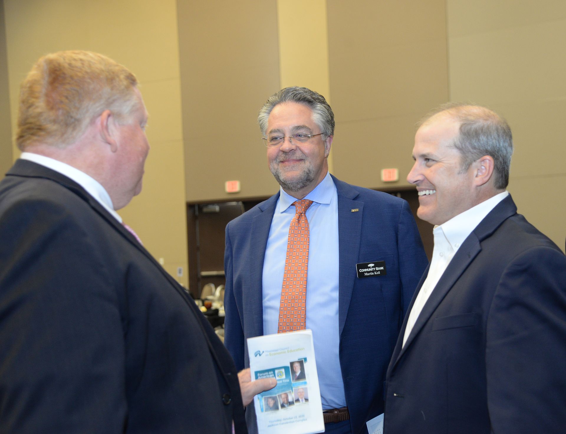 Three men in suits conversing in a large room. One holds printed material.