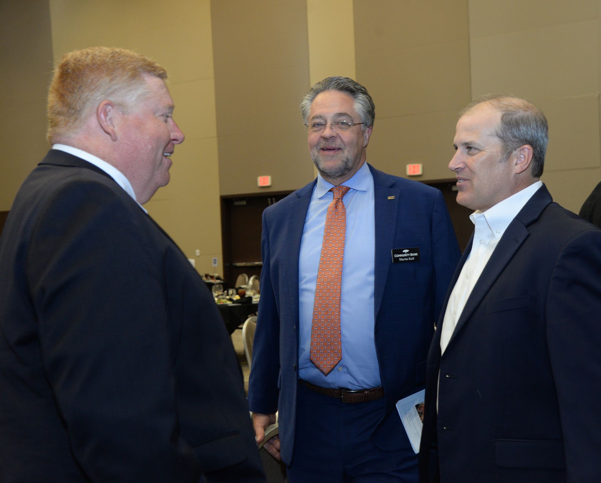 Three men in suits conversing indoors; one wears an orange tie.