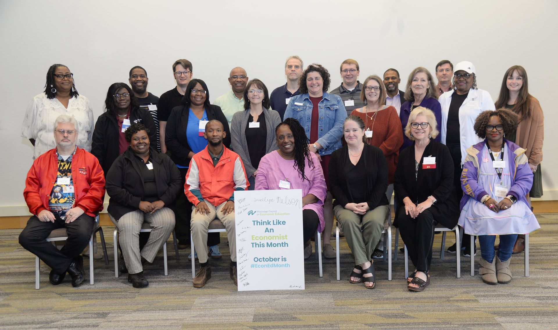 Group of people at an event pose with a sign. People in business casual attire in a brightly lit room.