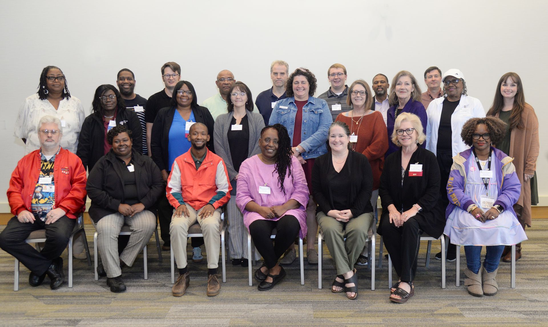 Group of people posing for a photo. They are seated and standing against a white wall. Some are wearing name tags.