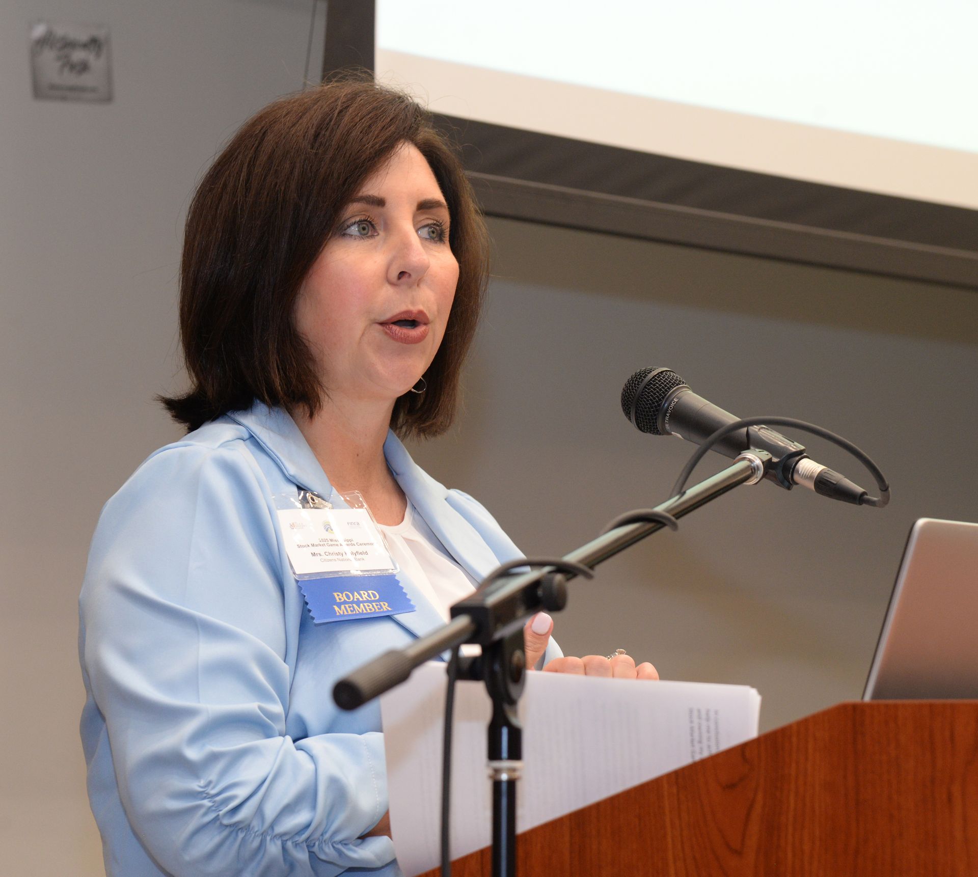 Woman speaking at a podium, wearing a light blue blazer, holding papers and speaking into a microphone.
