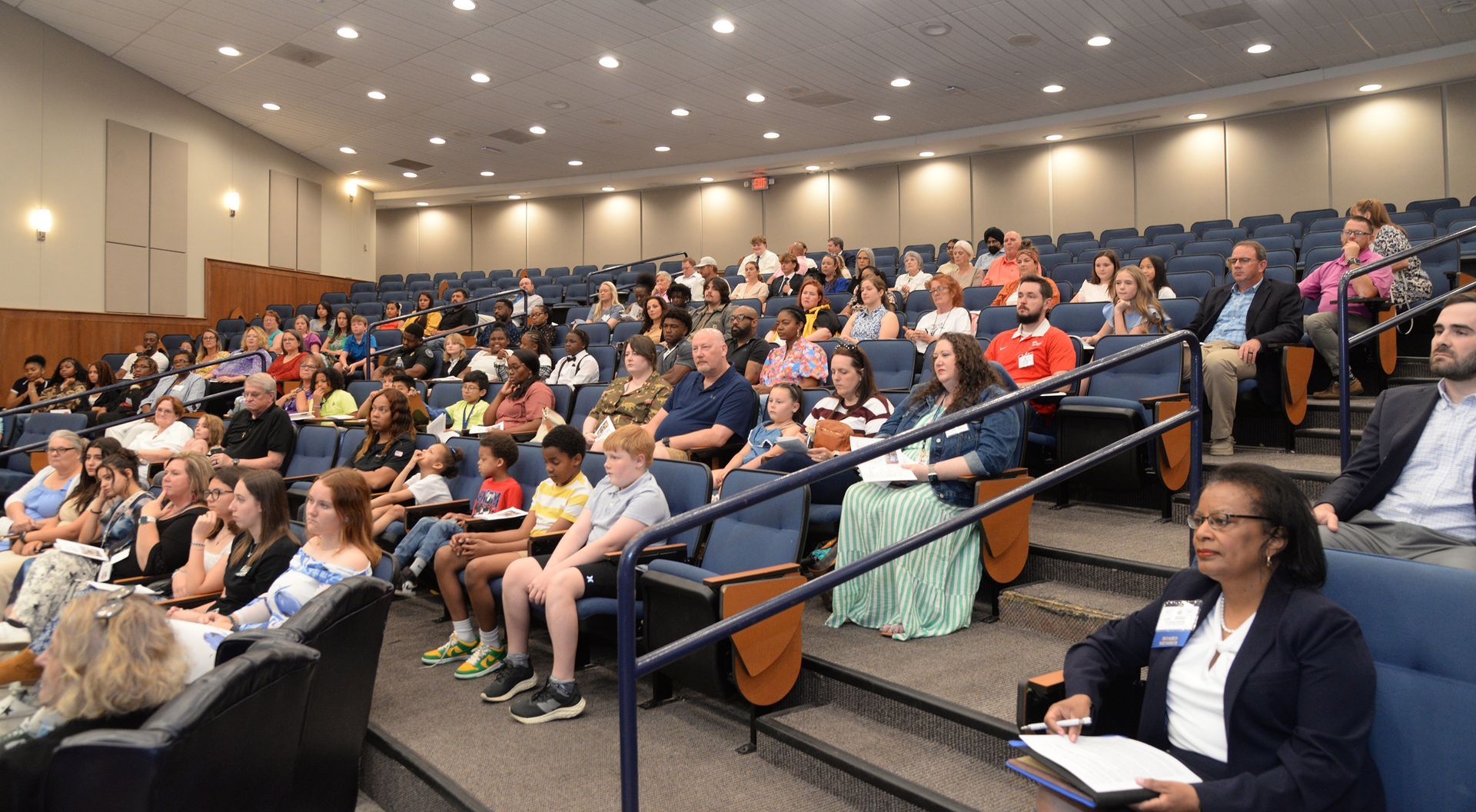 Audience in a tiered auditorium. People sit and listen; some hold notebooks. The room has blue seats and is brightly lit.