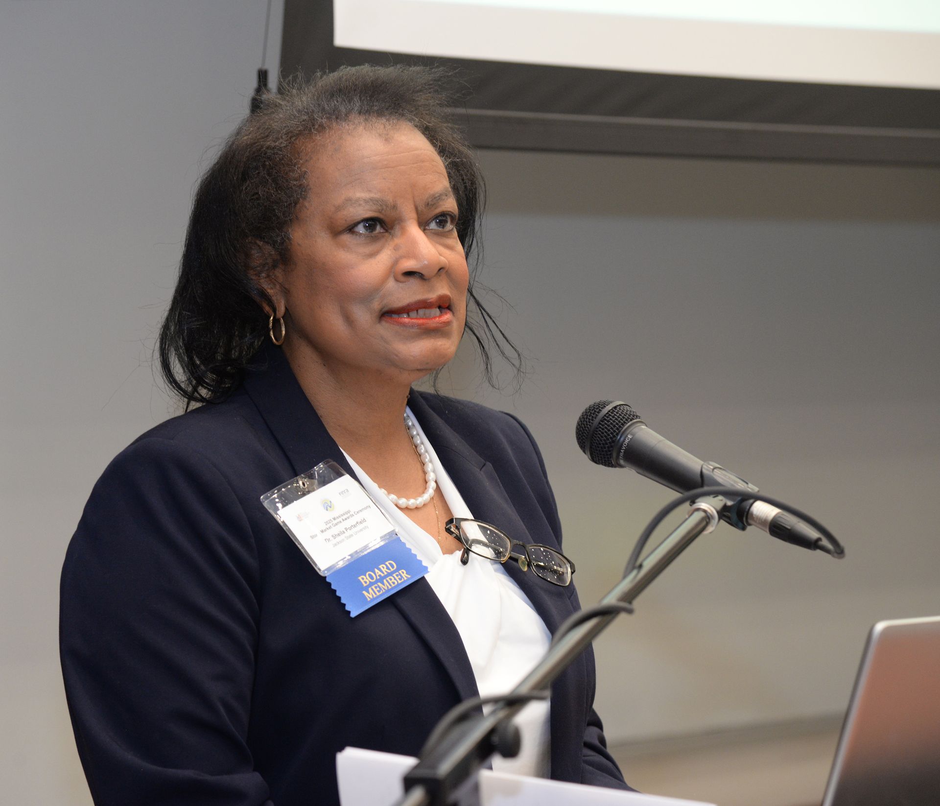 Woman speaking at a lectern with a microphone, wearing a blazer, in a conference setting.