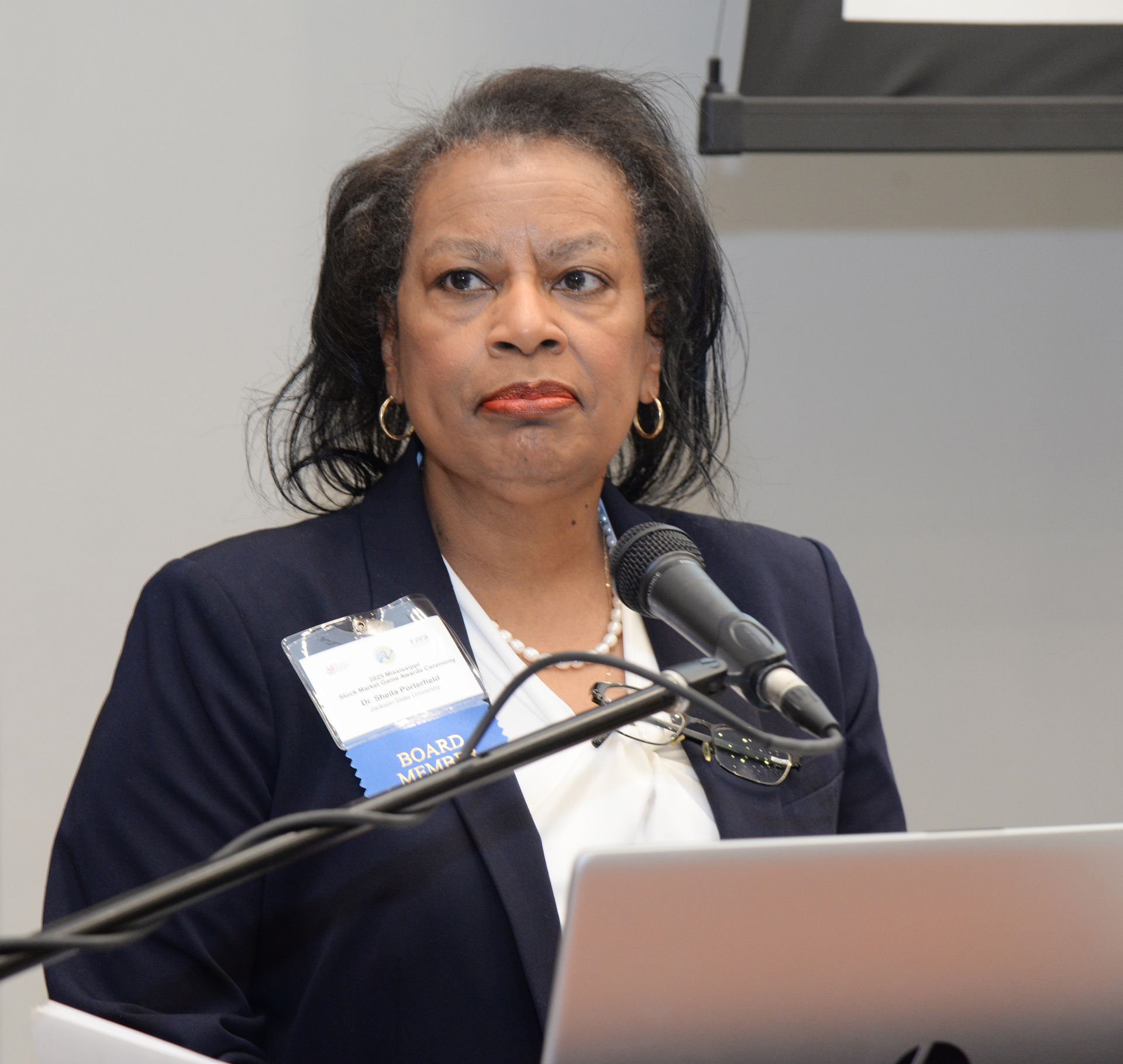 Woman speaking at a lectern, wearing a blue blazer and white blouse, holding a microphone.