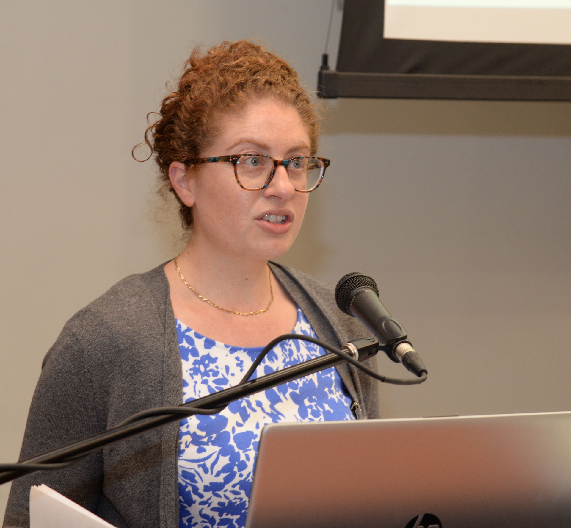 Woman speaking at a lectern, wearing glasses, blue and white floral top, gray cardigan. Microphone present.