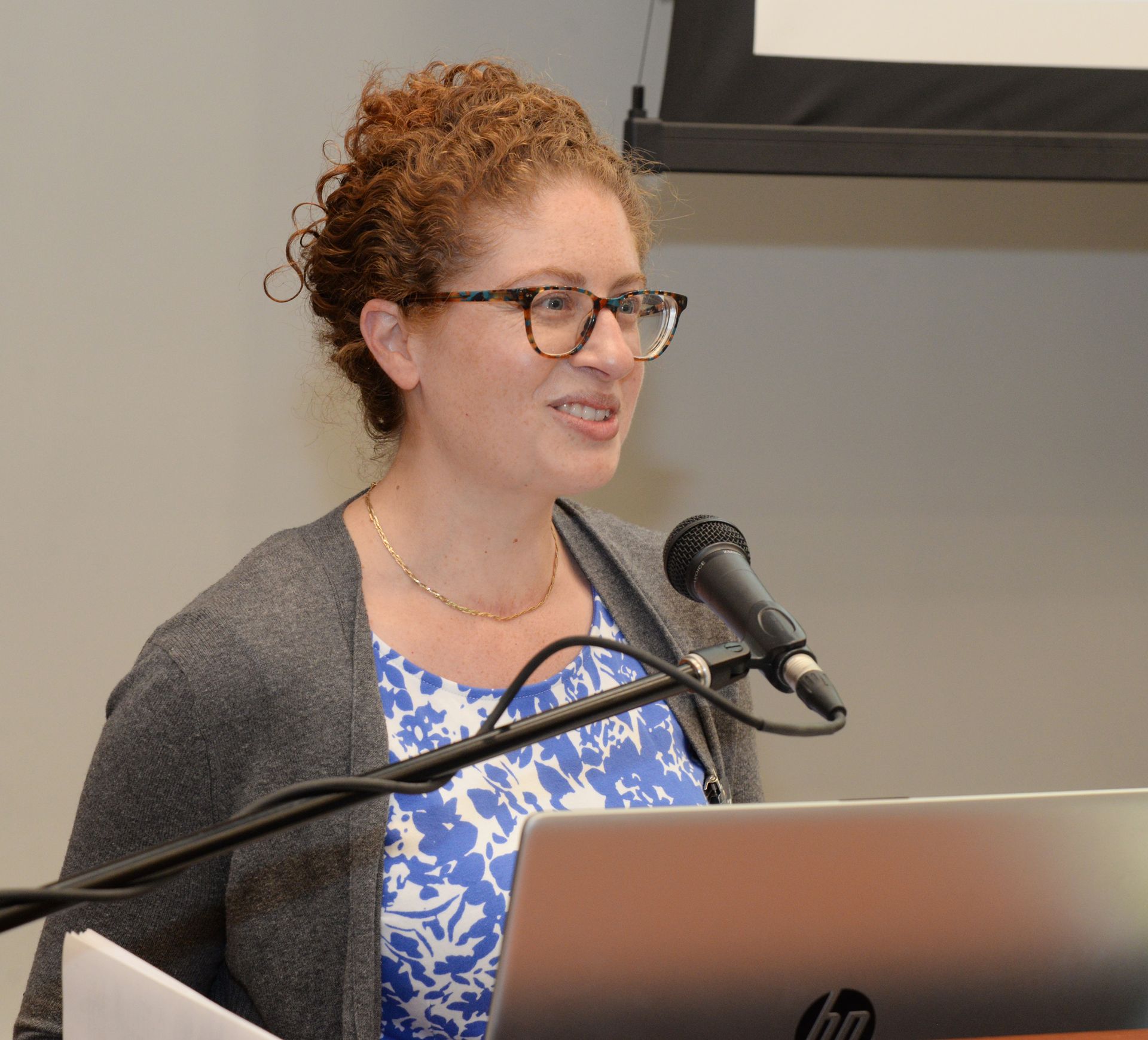 Woman speaking at a lectern, wearing glasses and a gray cardigan over a blue and white patterned shirt.
