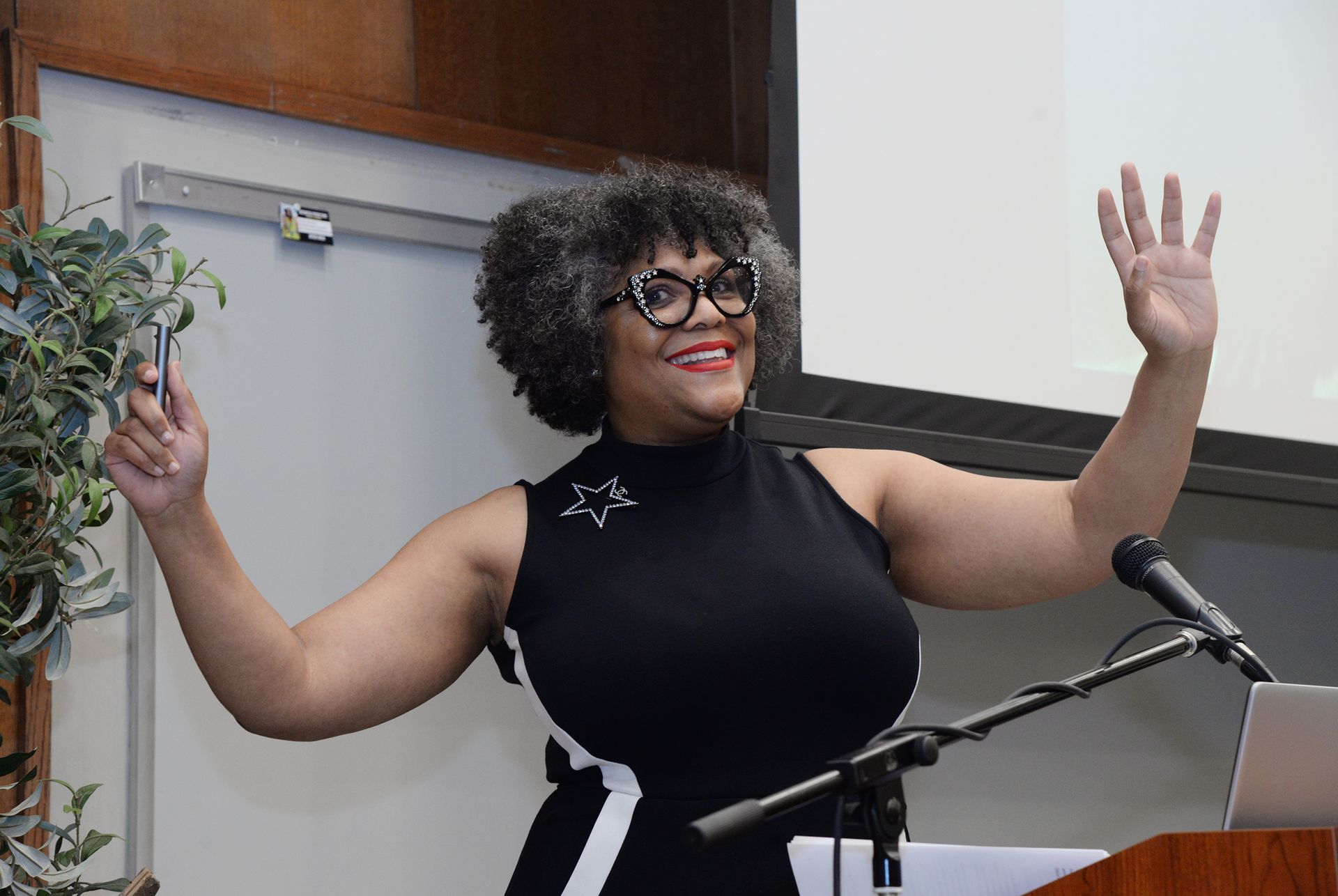 Woman with curly hair and glasses waves while speaking at a podium.