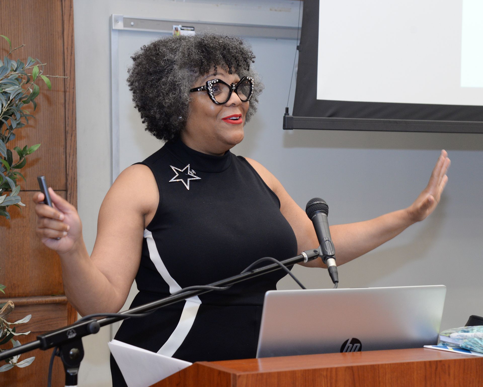 Woman presenting at a podium, gesturing with open hands, laptop, microphone, and a presentation screen in the background.