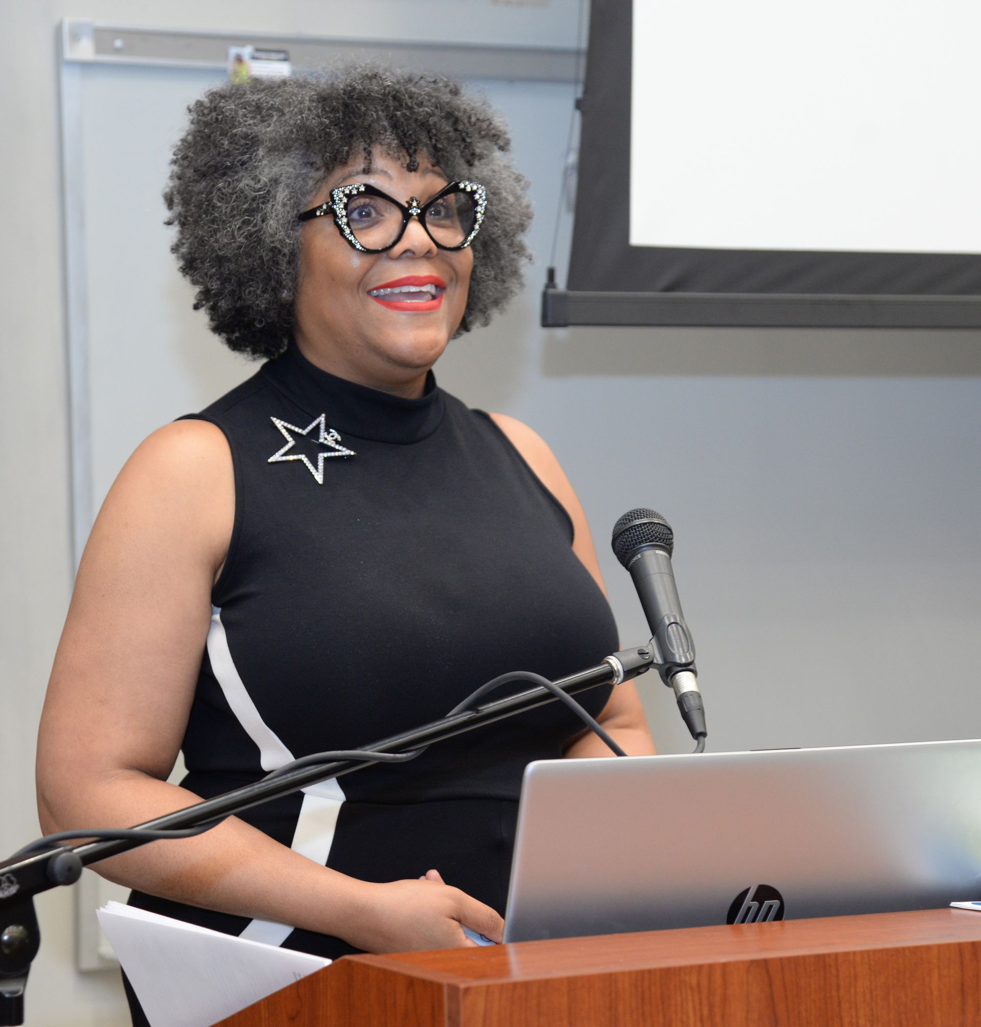 Woman speaking at a podium, wearing glasses, black dress with star pin, and silver hair. Microphone and laptop present.