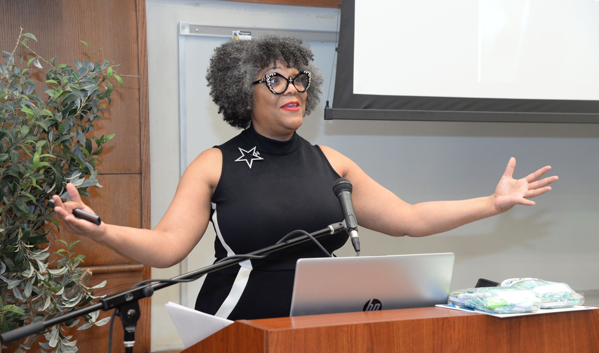 Woman speaking at a lectern with open arms, in front of a screen. Wearing glasses and a black dress.