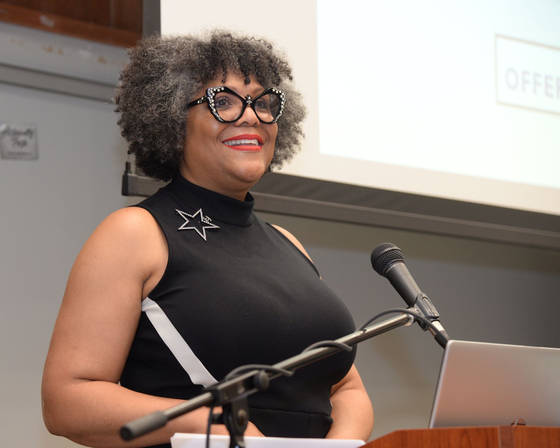 Woman at a lectern, speaking, with microphone and laptop. She wears glasses and a black top with star.