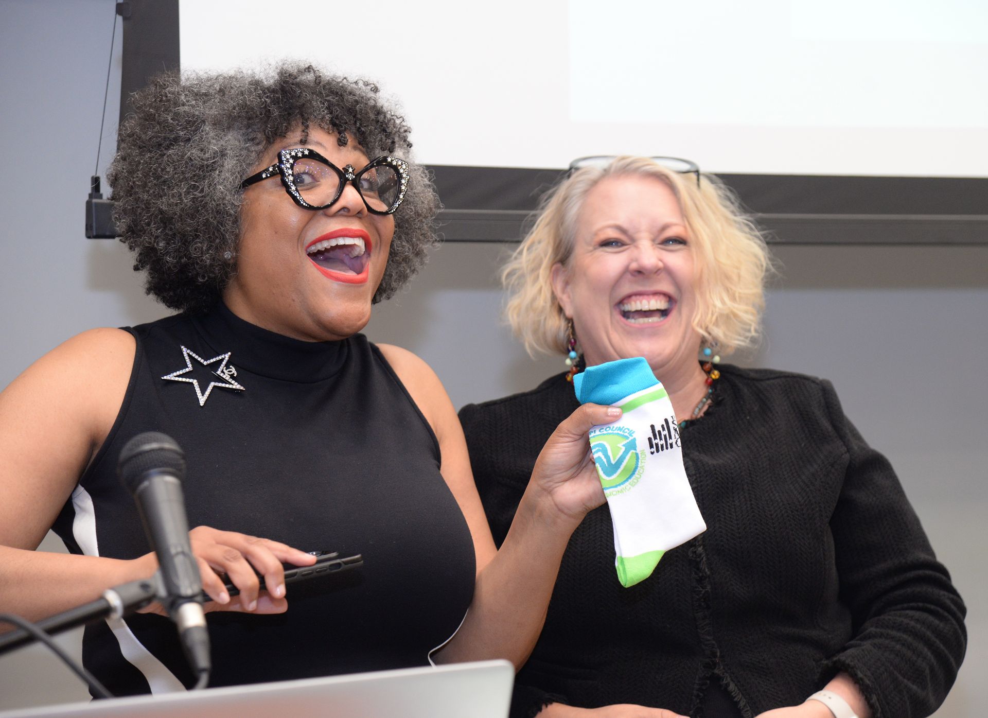 Two women laughing, one holding a sock, in front of a projection screen.