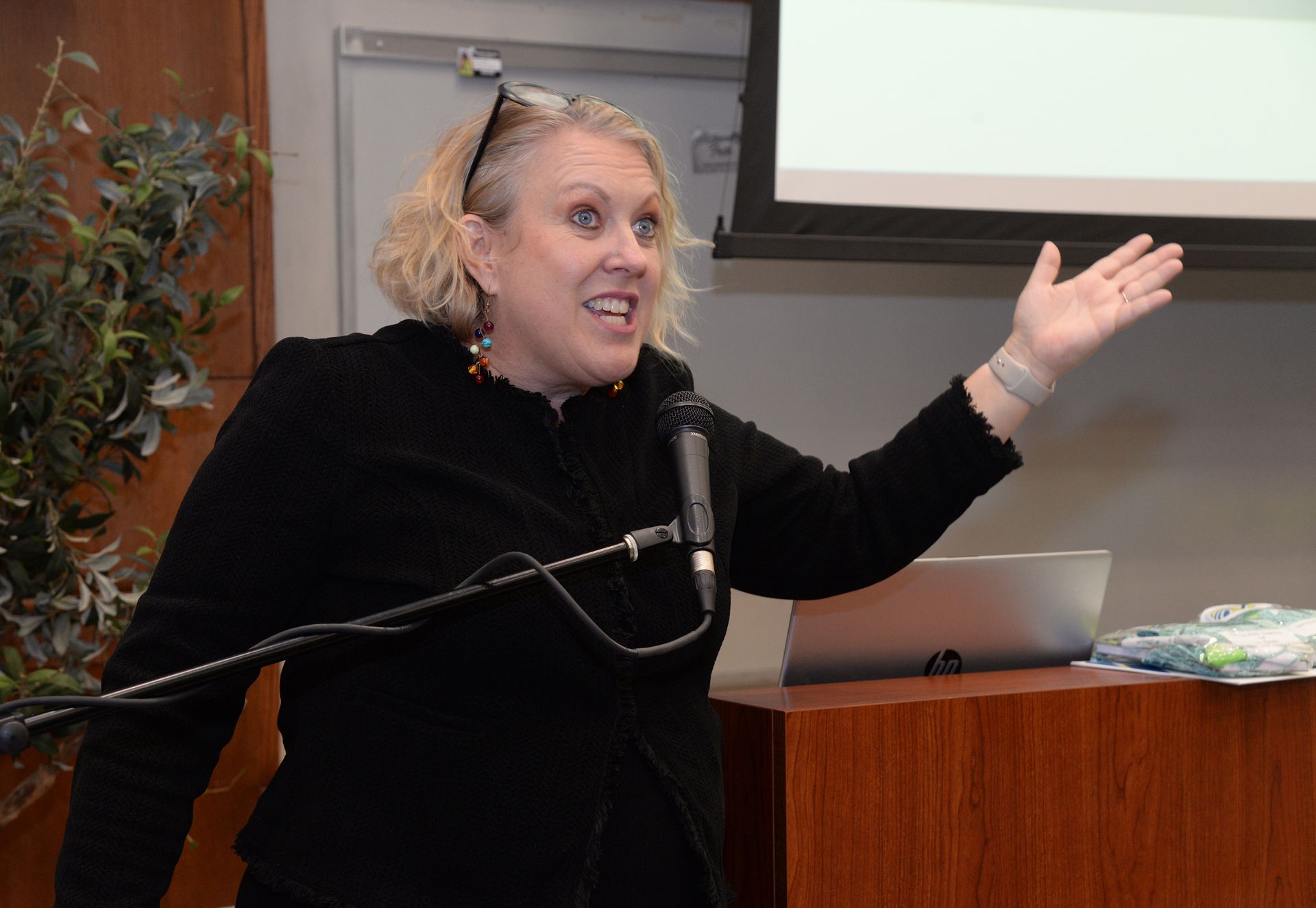 Woman gesturing animatedly at a podium, speaking into a microphone. Room setting with laptop and presentation screen.