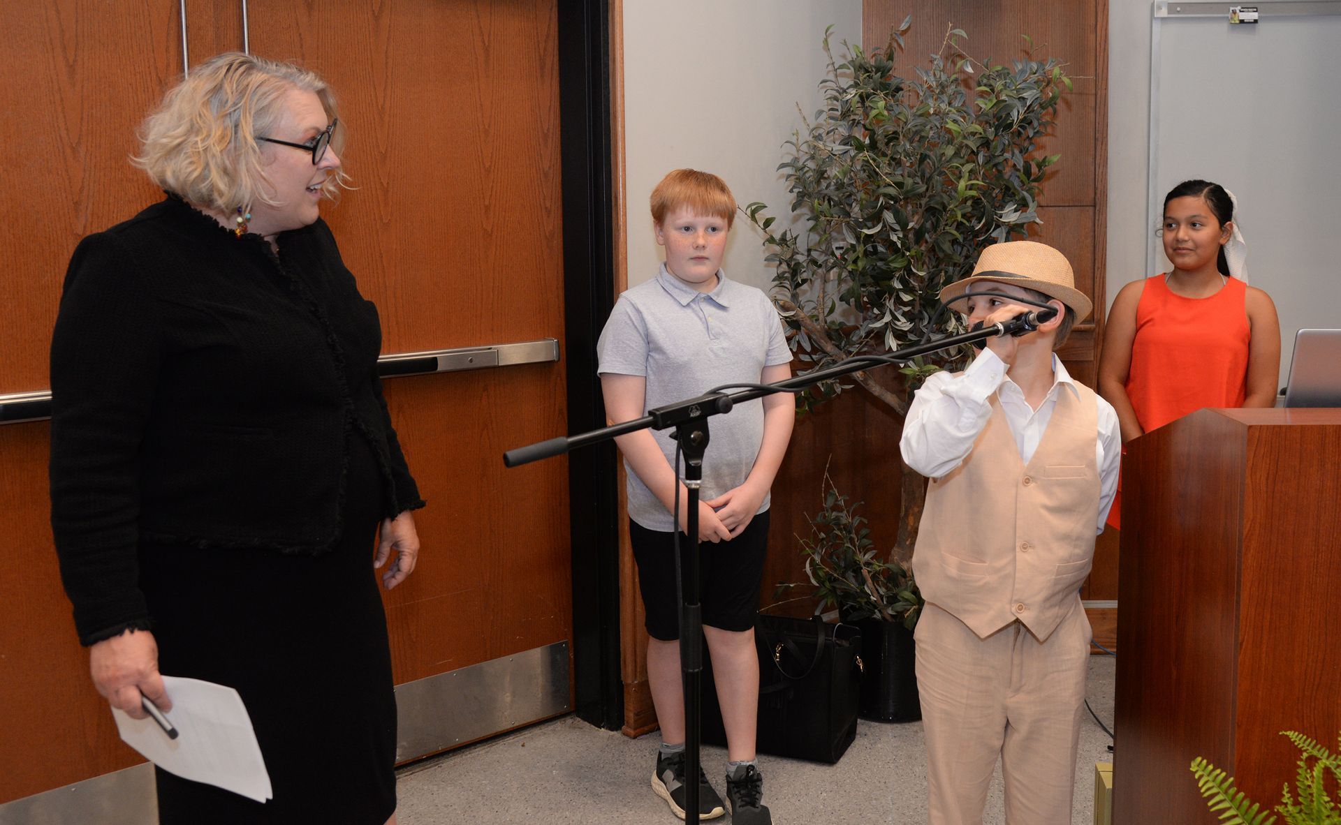 A boy in tan suit speaks into a microphone as others observe.