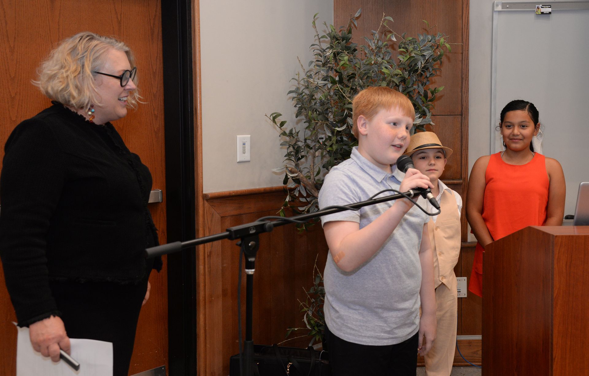 Woman and children at a podium. Boy speaks into a microphone. Another child stands next to him.