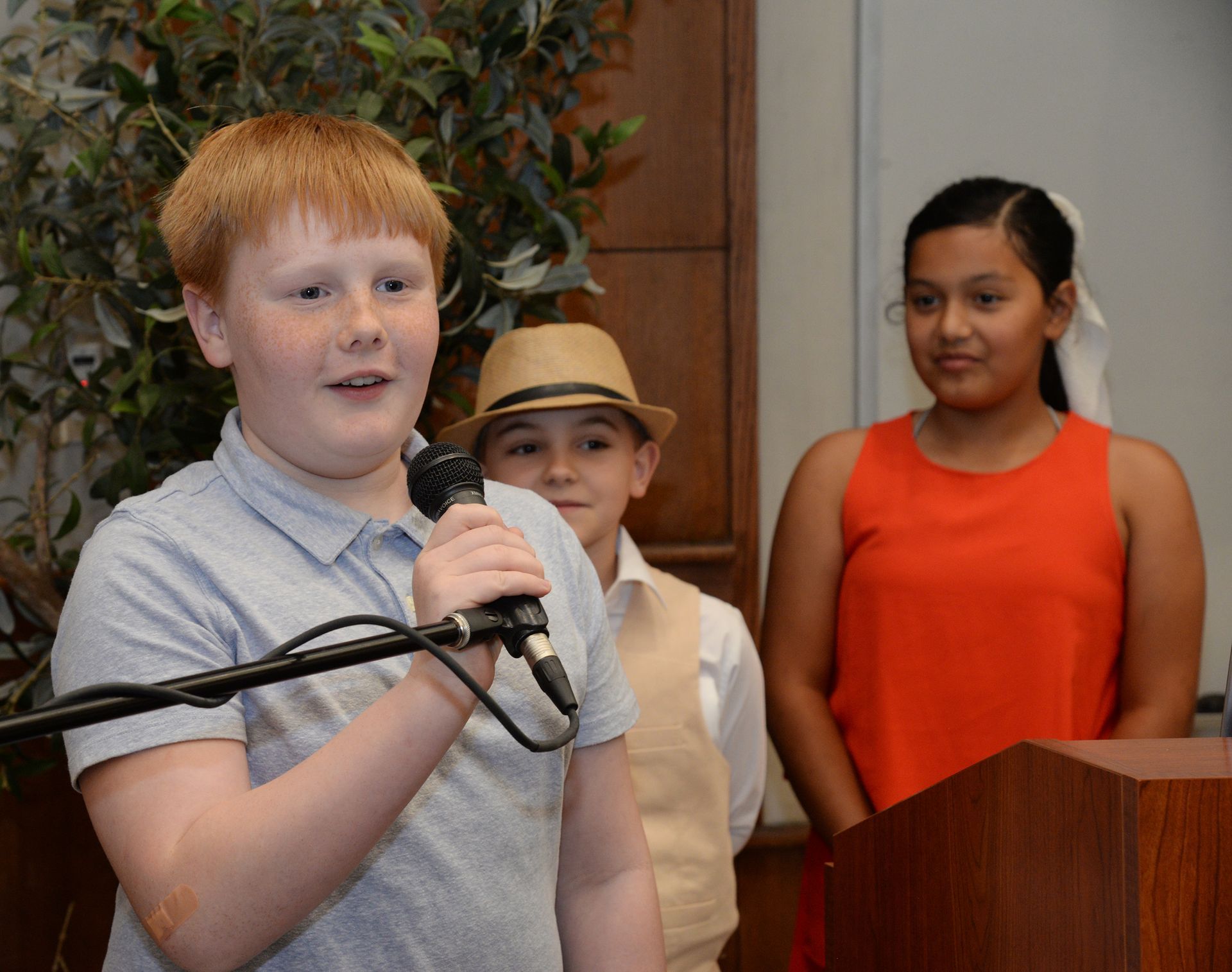 Boy speaking into microphone at a podium, two children in background.