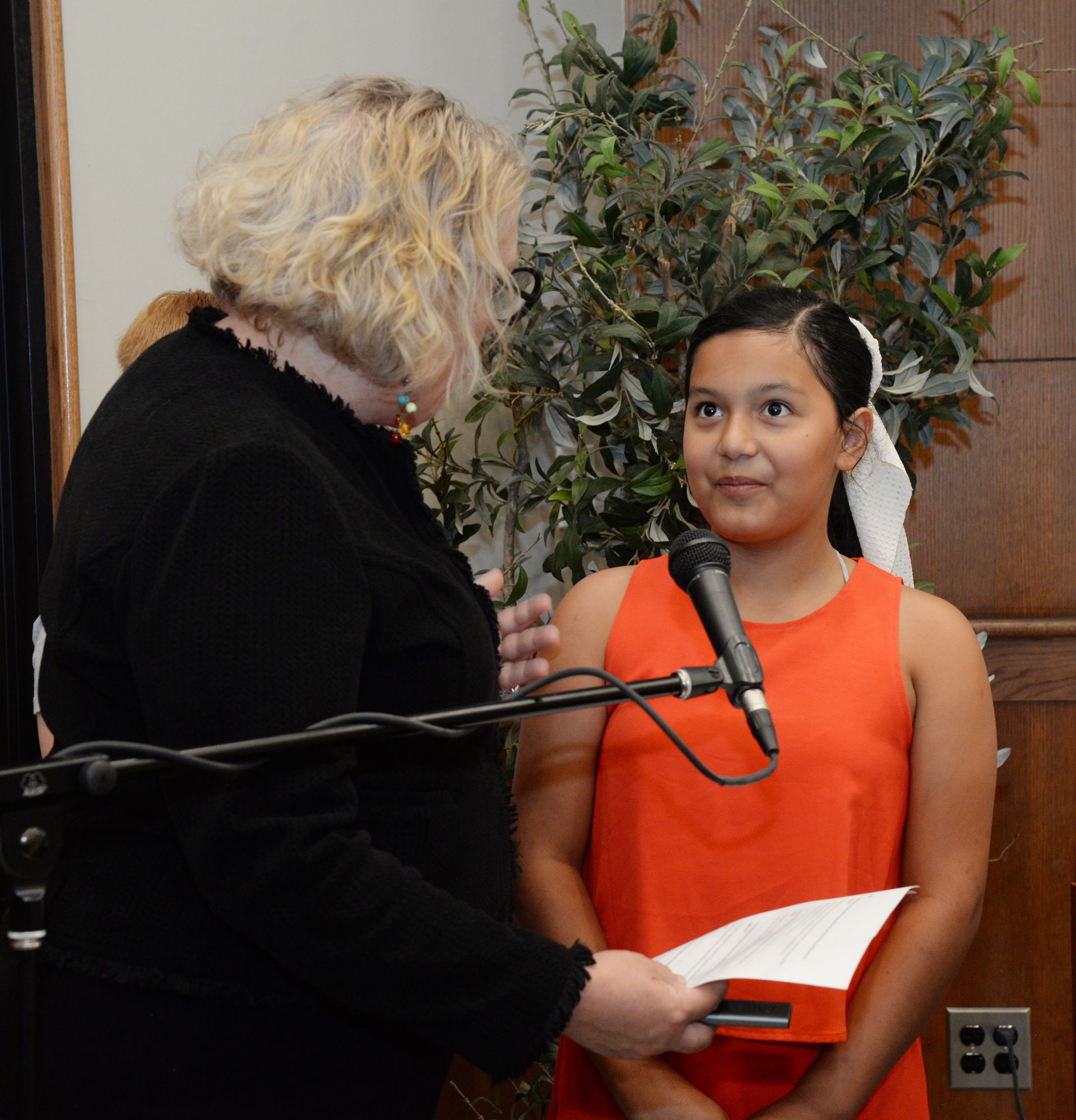 Woman in black jacket speaks to girl in orange dress holding paper, near microphone and greenery.