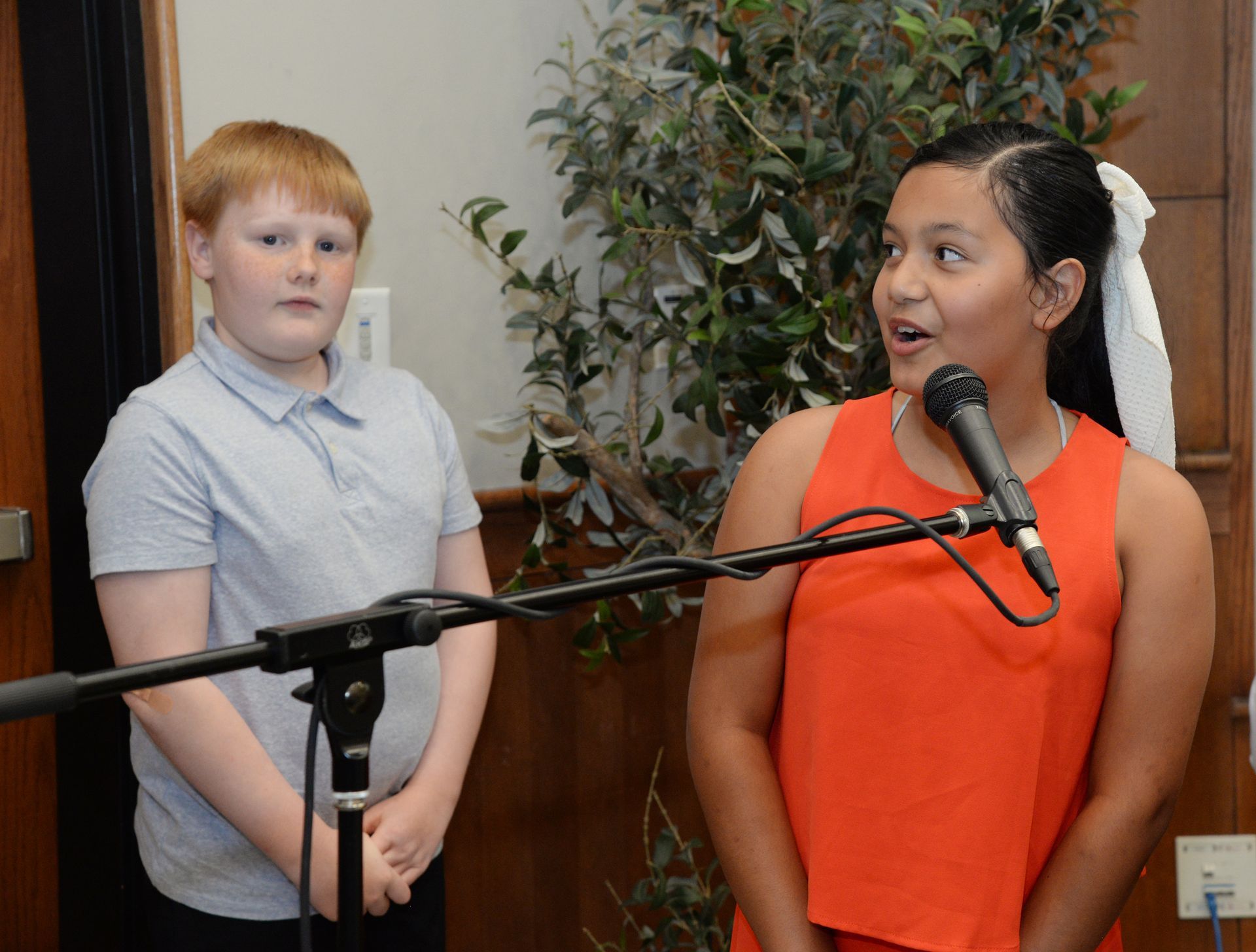 Two children speaking into a microphone. One wears an orange top, the other a grey shirt. Indoor setting.