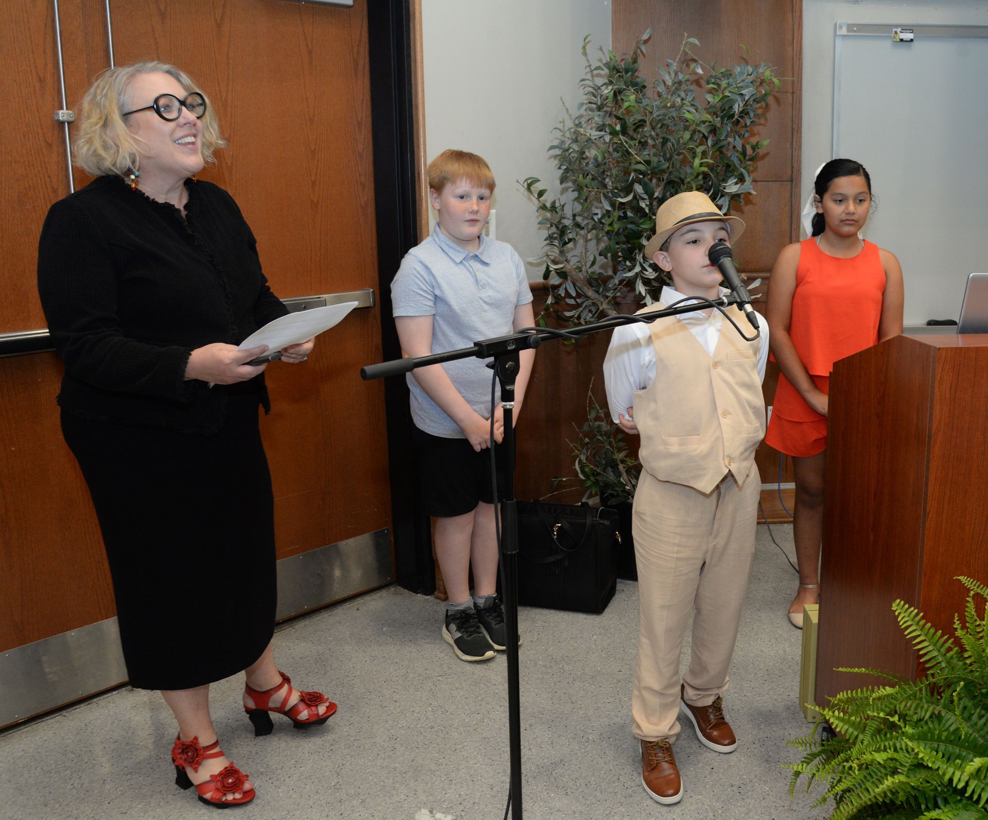 Woman, children at podium. Boy with hat speaks into microphone. Others stand nearby.